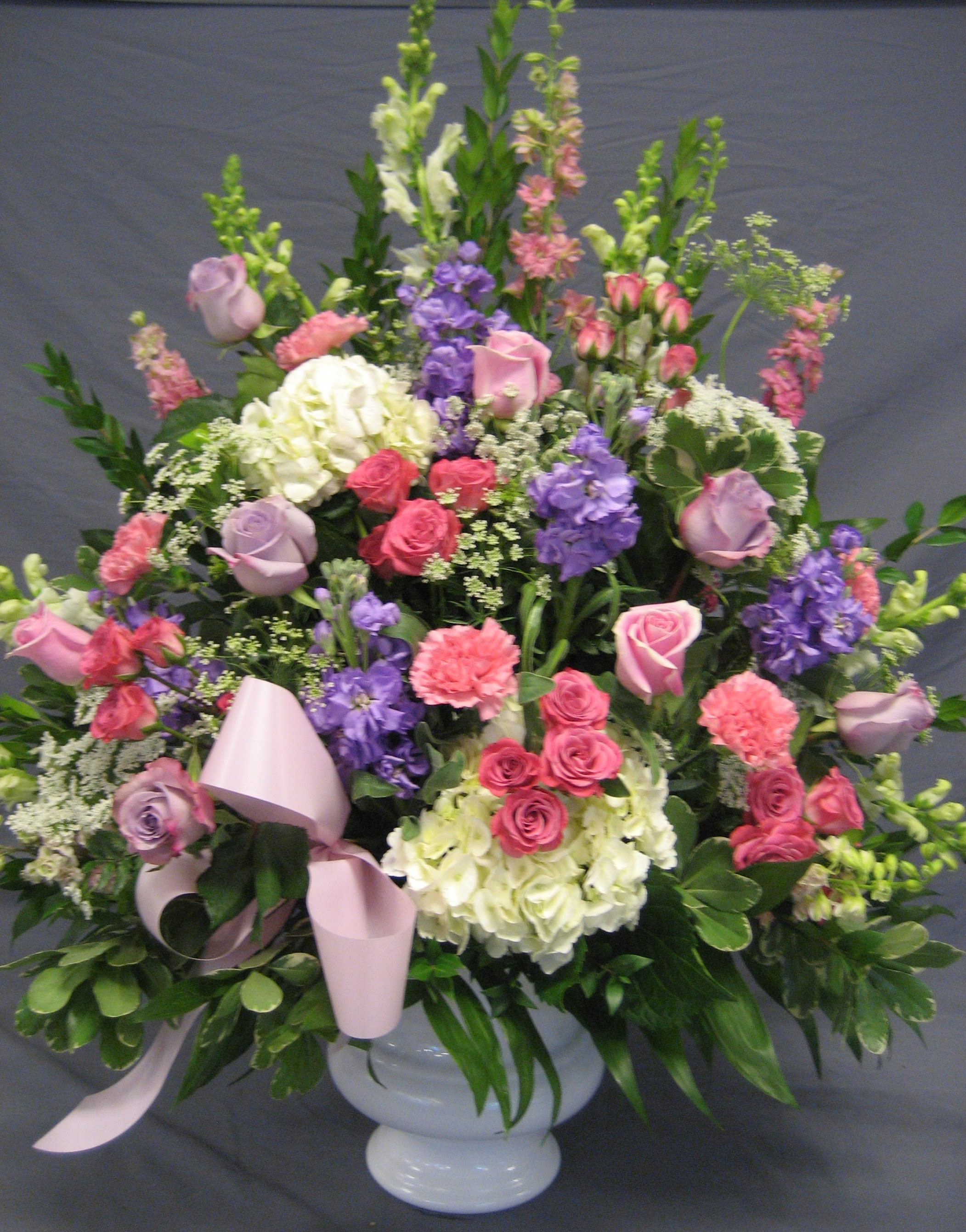 Large mixed floral arrangement in a white pedestal vase with pink ribbon