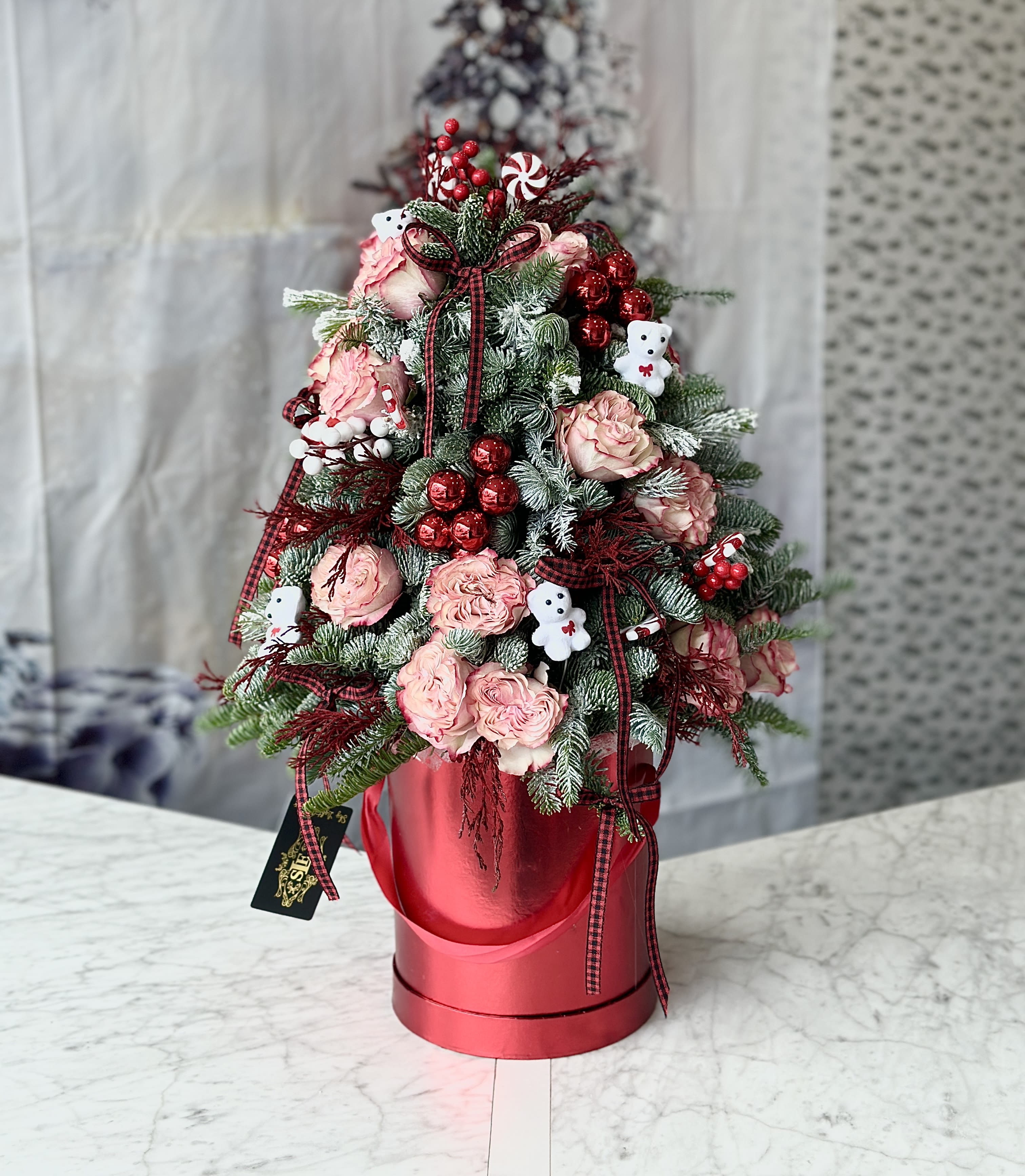 Pink roses and holiday greenery arranged in a red hatbox with ribbon and teddy bear picks