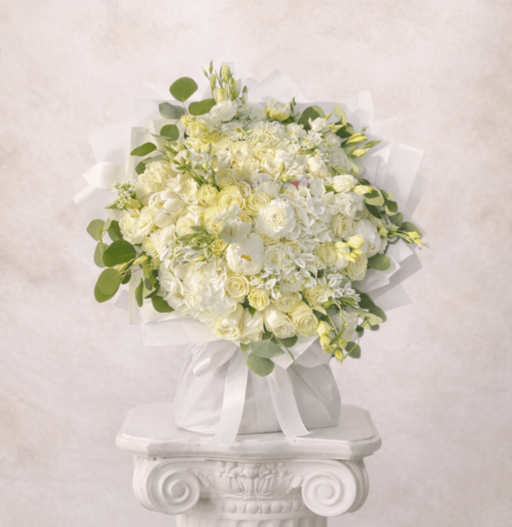 Round bouquet of white and pale yellow flowers wrapped in white fabric on a white pedestal.