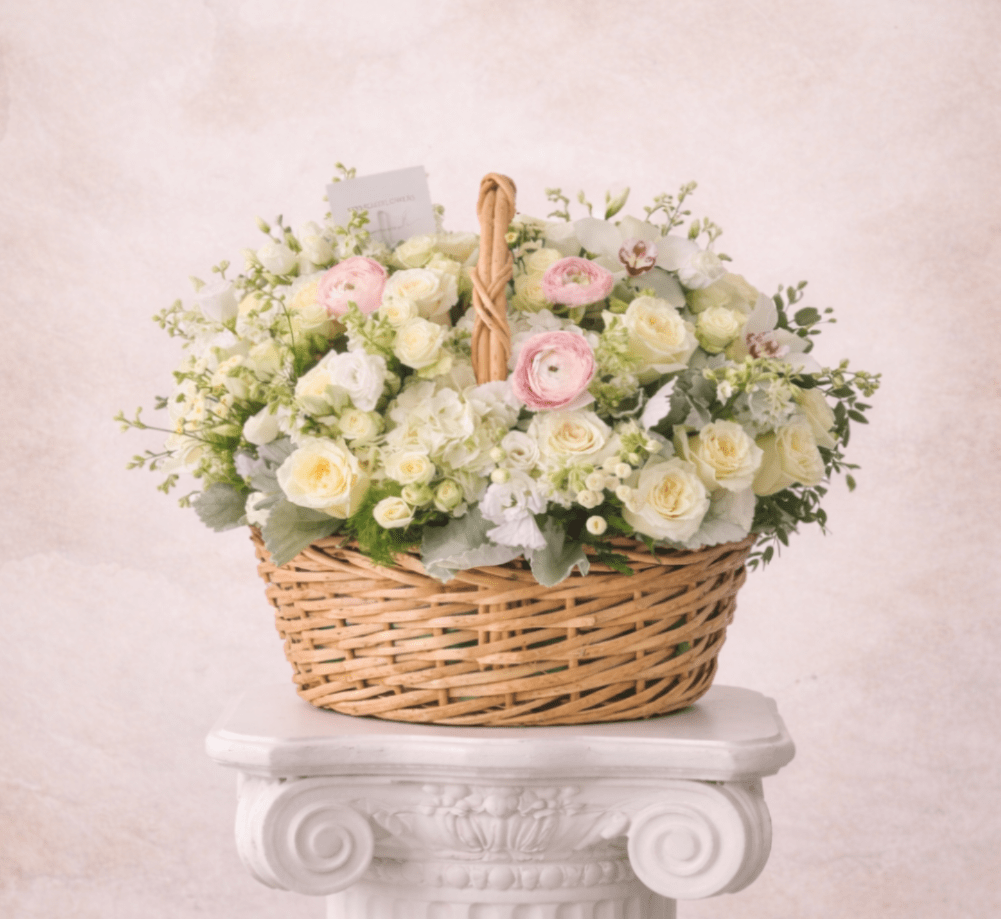 Basket arrangement of cream roses with soft pink ranunculus and white blooms on a white pedestal.