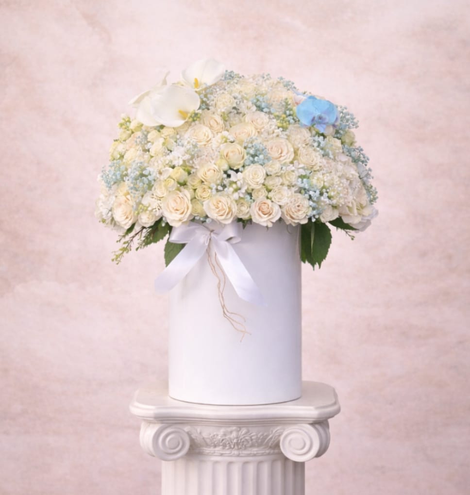 Round arrangement of ivory spray roses with white and blue accents in a tall white cylinder box.