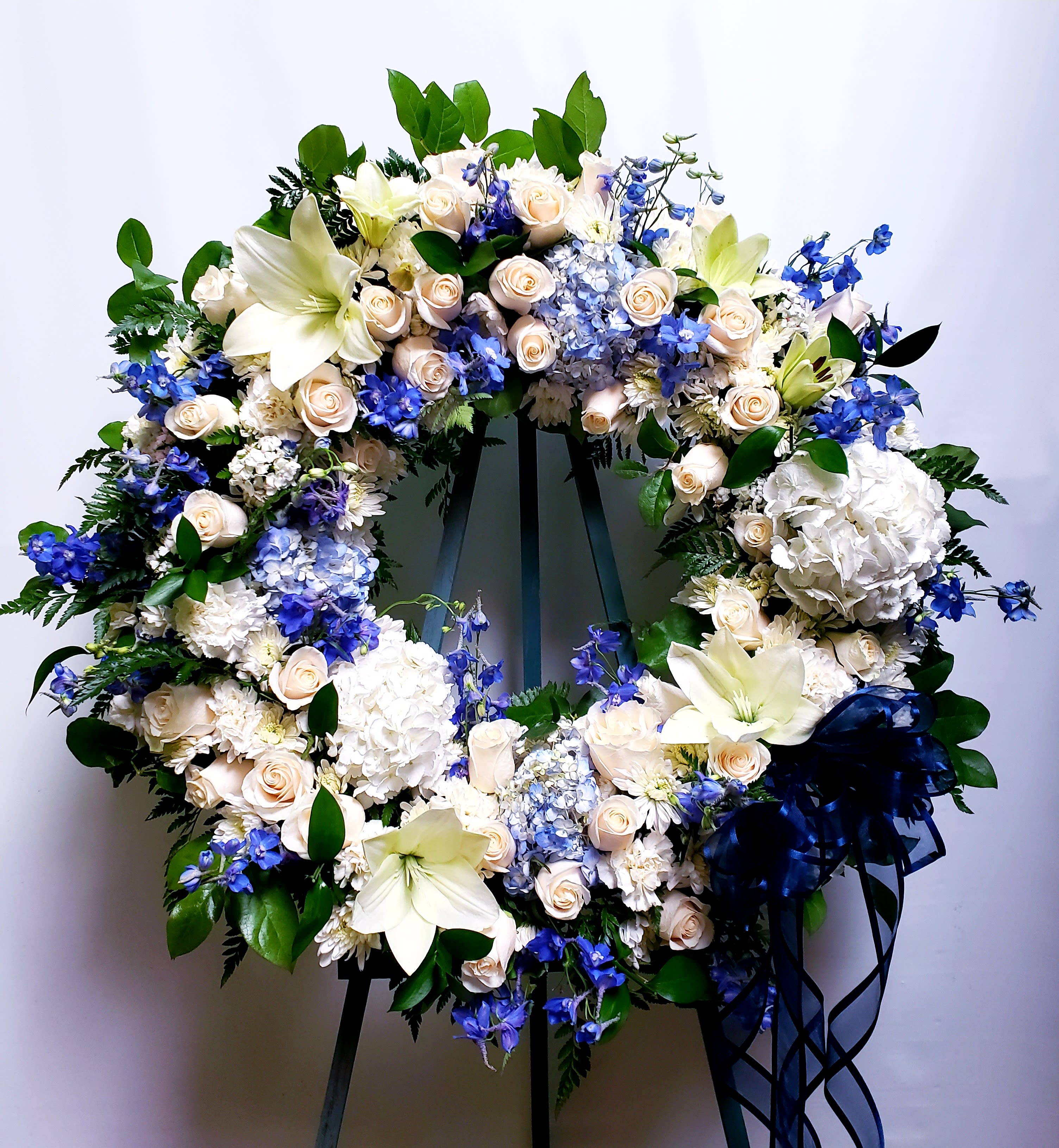 Large floral wreath with white lilies, cream roses, blue flowers, and a navy ribbon