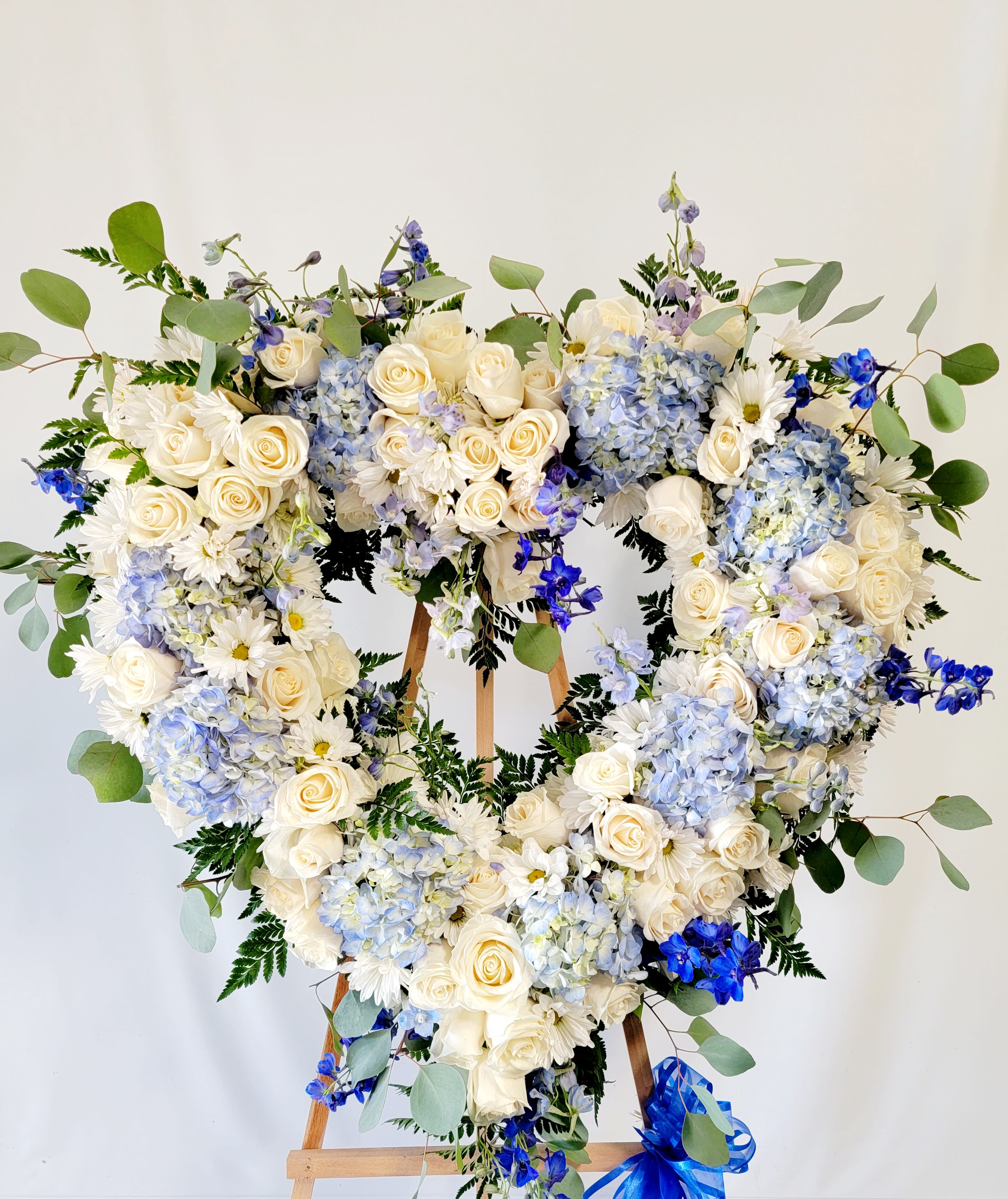 Heart-shaped floral wreath with white roses, blue hydrangeas, and daisies on an easel