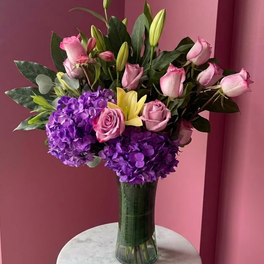Pink roses, purple hydrangeas, and yellow lilies in a tall glass vase
