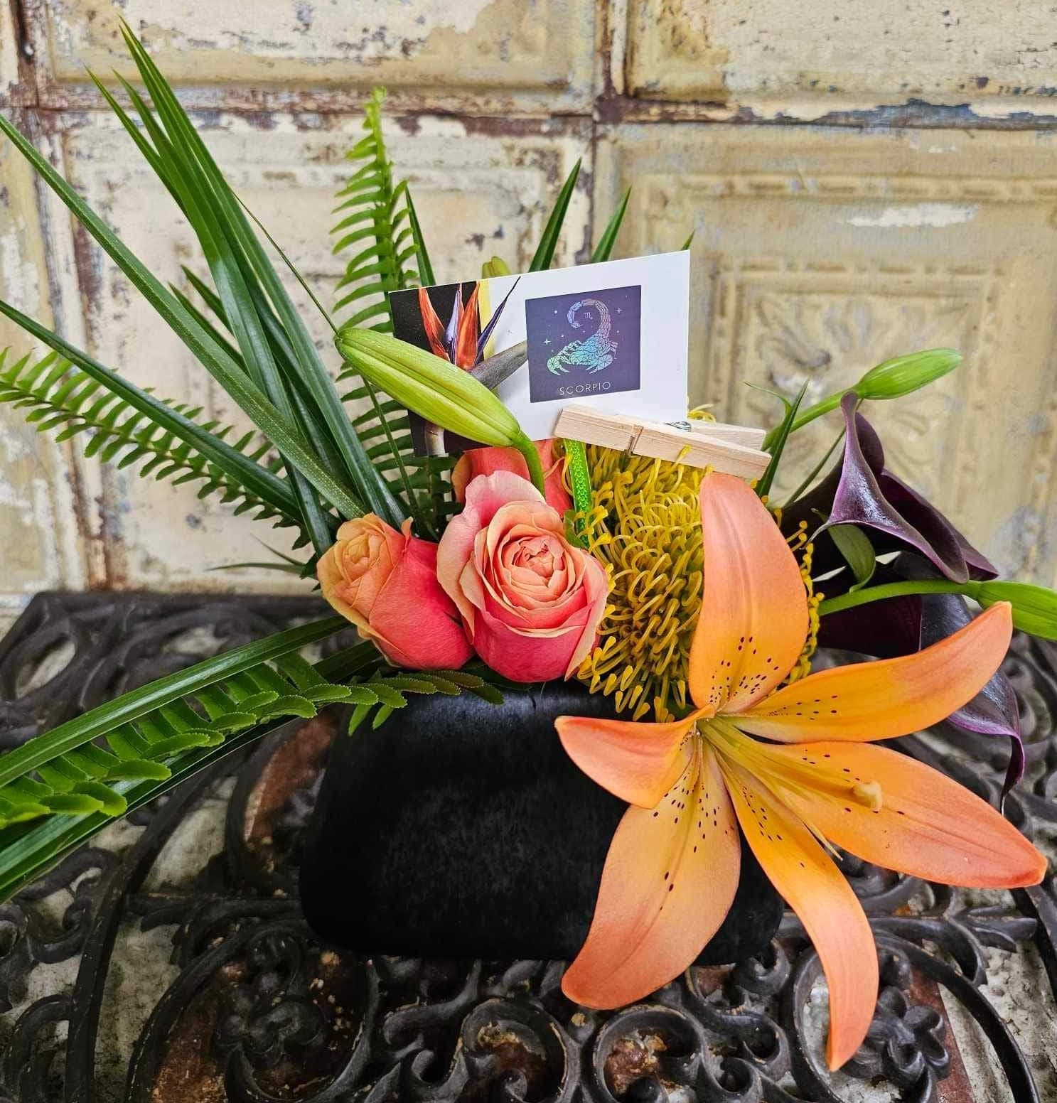 Tropical floral arrangement with roses, lilies, and a Scorpio card in a black container