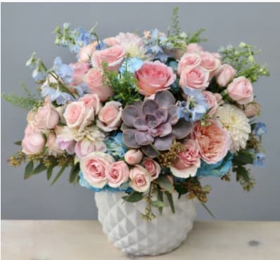 Pink roses and blue flowers arranged in a white vase with a succulent center