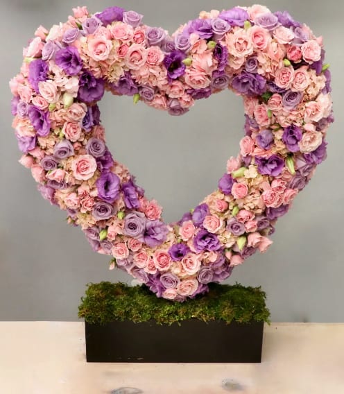 Large heart-shaped arrangement of pink and purple flowers on a moss-covered black rectangular base