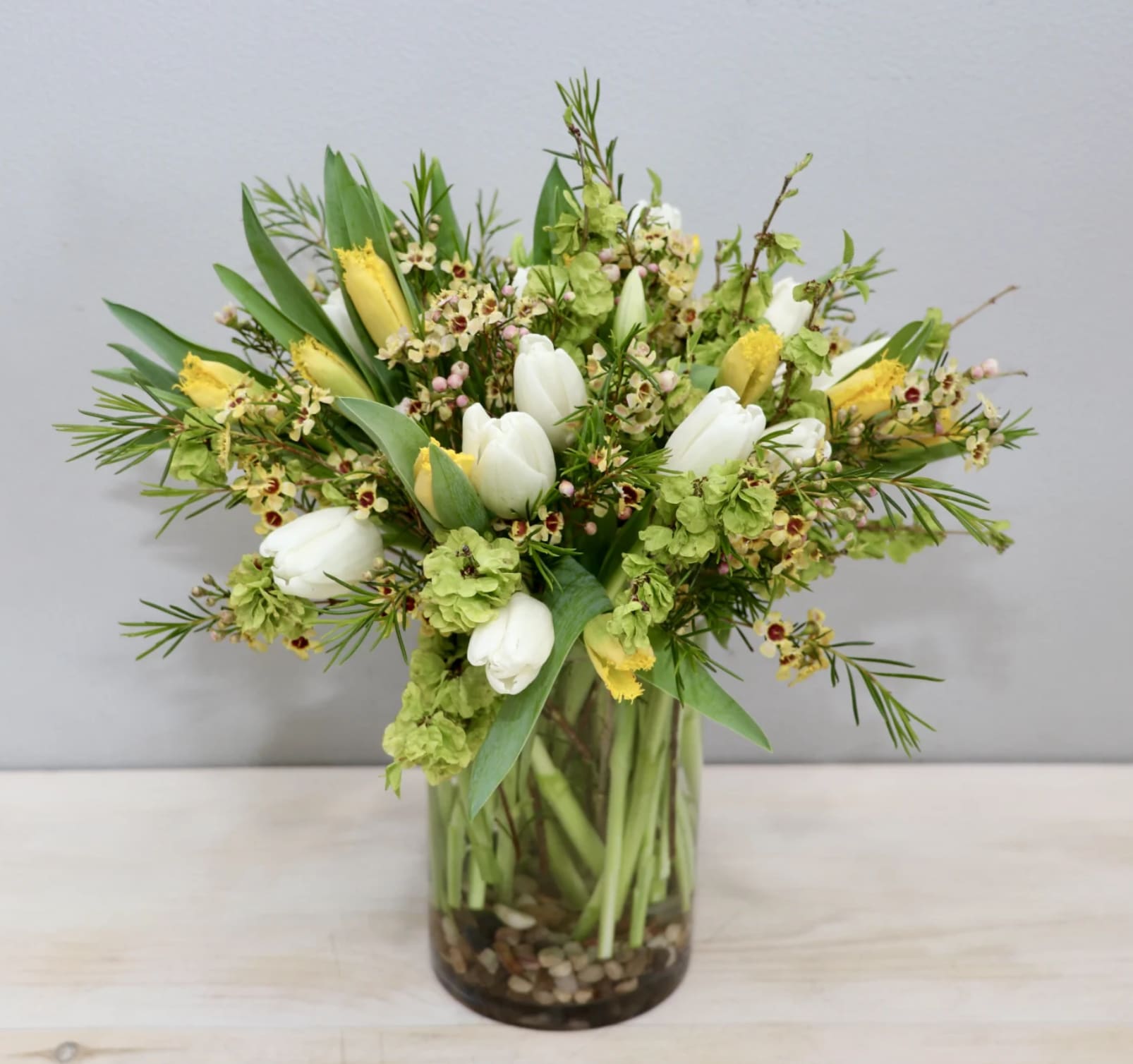Arrangement of white and yellow tulips with green blooms in a clear glass vase with stones