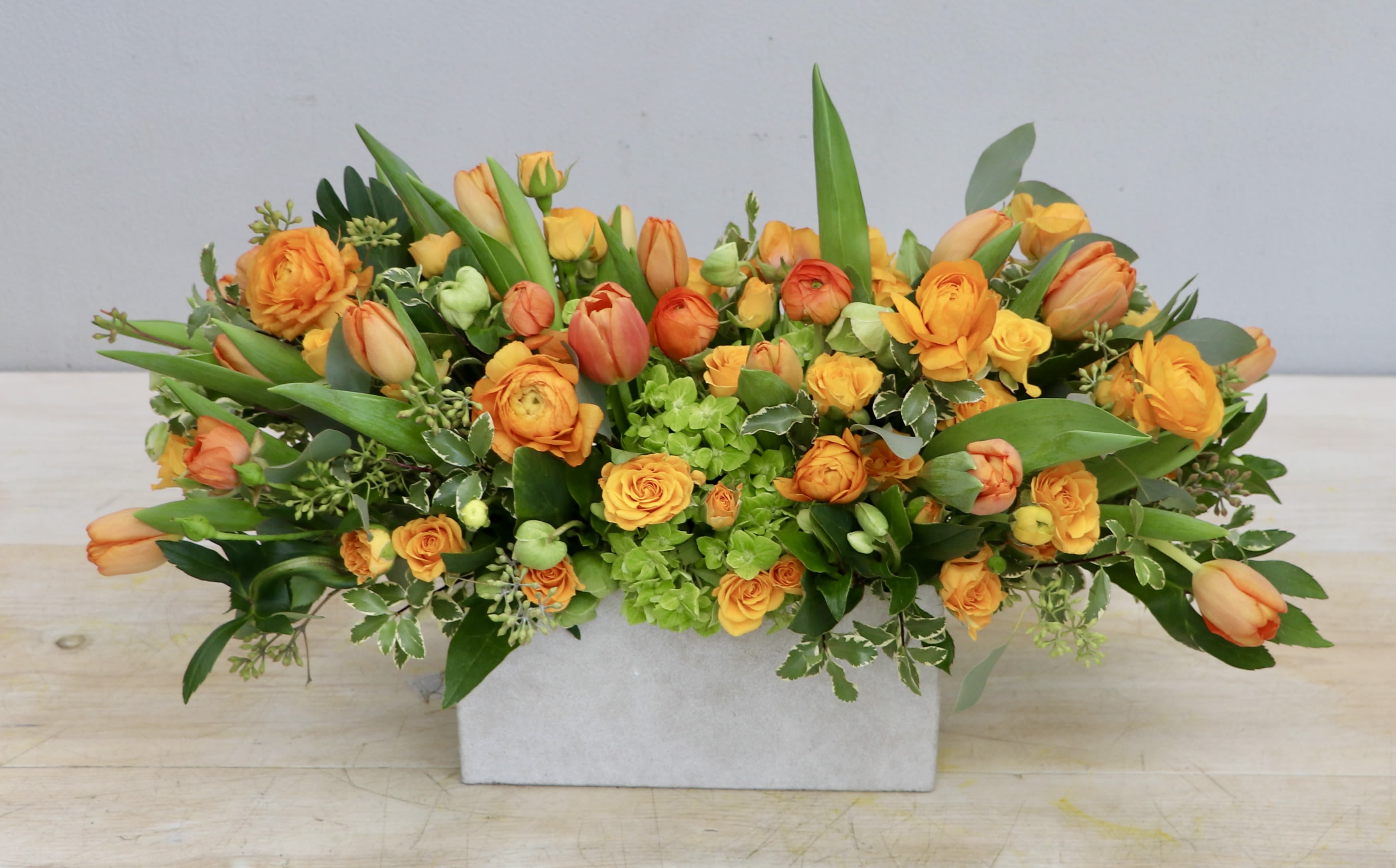 Orange Sunrise - WeHo - Orange Sunrise showcases orange ranunculus, tulips, and spray roses in a bed of seasonal greenery. This arrangment is about 14" across for the standard size and gets larger with each upgrade!