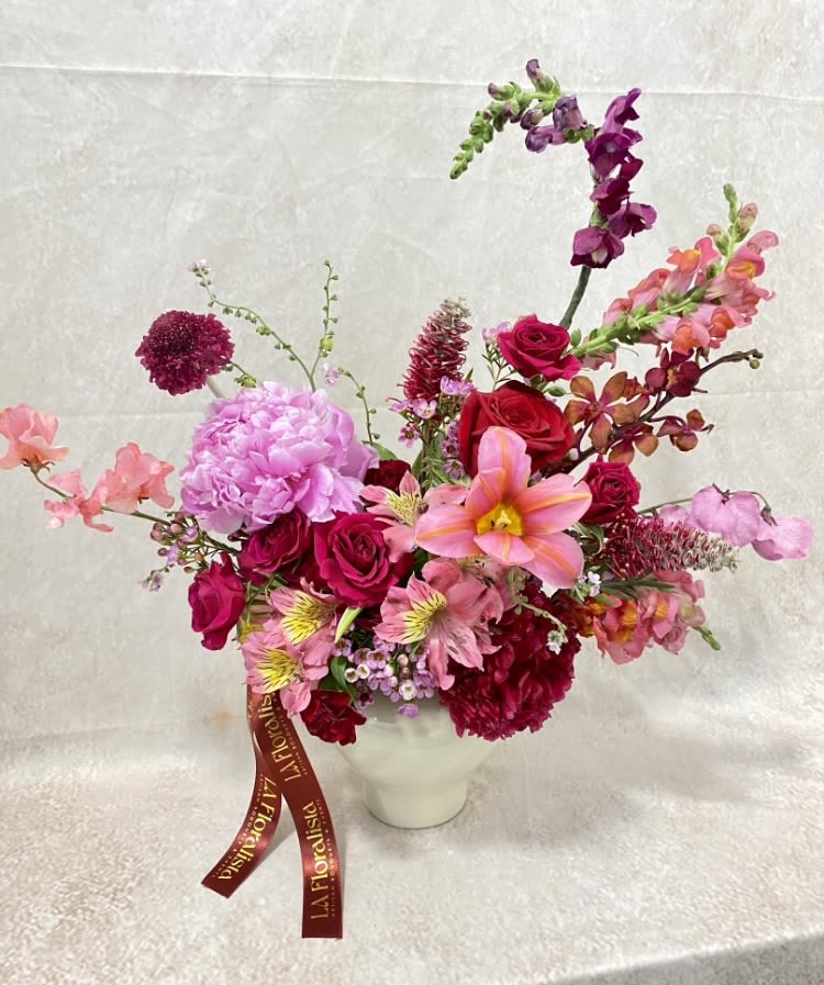 Pink and red mixed flower arrangement in a white vase with a ribbon