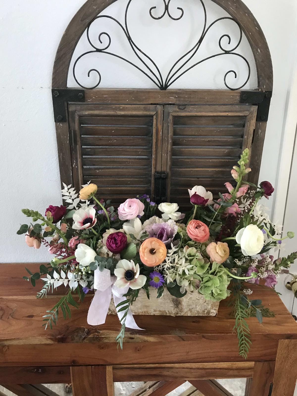 Low rustic centerpiece of mixed pastel ranunculus, anemones, and other blooms in a rectangular container with ribbon