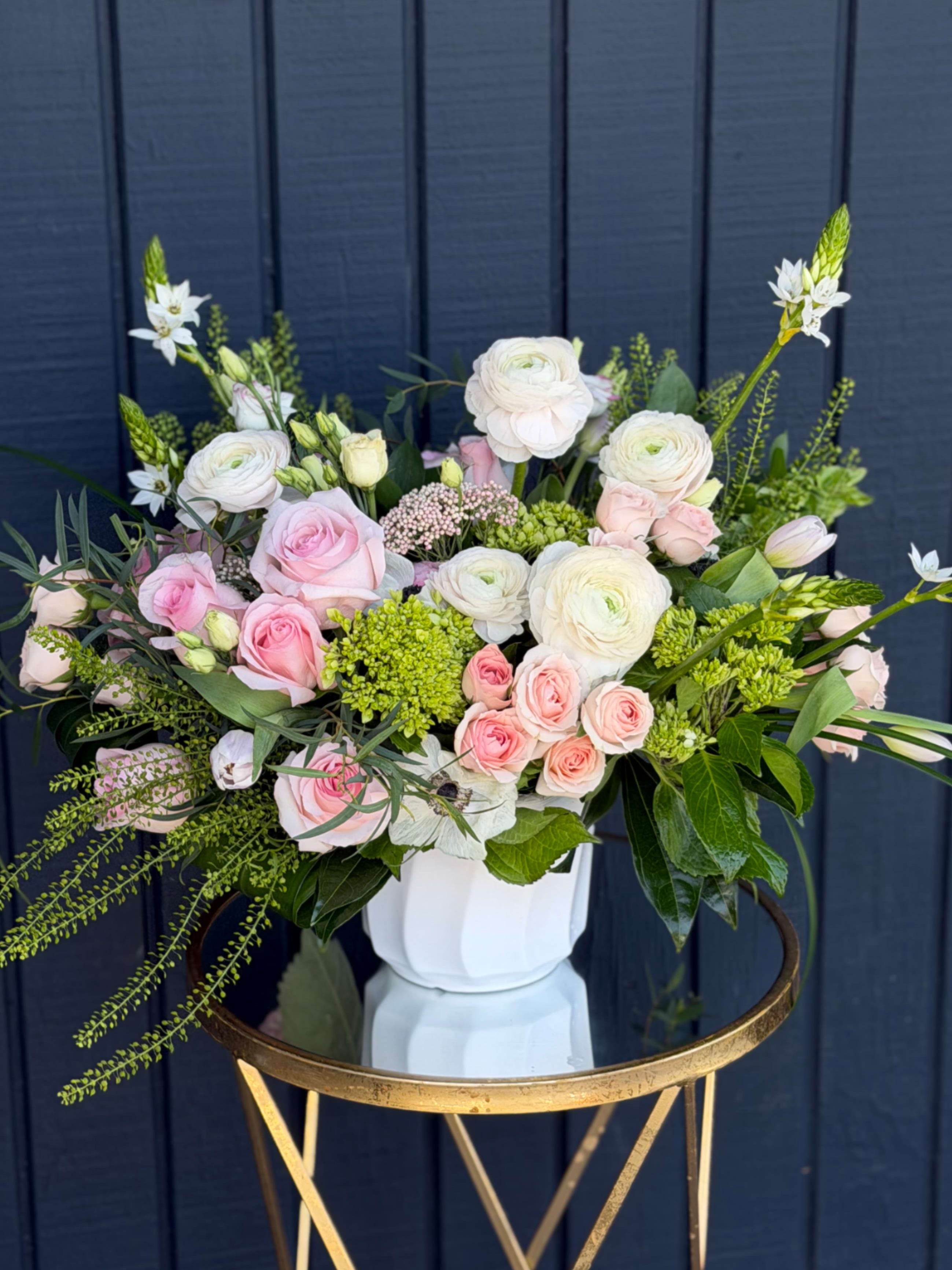 Fairytale garden  - Soft blush roses and creamy white ranunculus mingle with clusters of pink spray roses in a rounded, abundant Fairytale garden arrangement-perfect as a birthday bouquet, anniversary flowers, or a just-because gift that feels gently romantic. The soft pinks and bright whites read as light and airy, while fresh green accents and a few taller white blooms add movement so the person you're thinking of will feel both delighted and truly seen.  Arranged in a clean white ceramic vase, the composition combines garden-style looseness with modern polish-rounded blooms at the center, airy textural greens for contrast, and petite spray roses that lend a delicate, layered look. The vase gives it a neat silhouette for tabletops or a bedside, and the color palette makes this equally right for spring celebrations or everyday moments when you want to brighten someone's day.  This is the kind of gift that says "I'm thinking of you" with soft romance; ideal for moms, partners, friends, or anyone who appreciates a tender, elegant floral statement. Size and vase are as shown-no extra assembly required; swap water every 2-3 days and trim stems for the longest freshness.