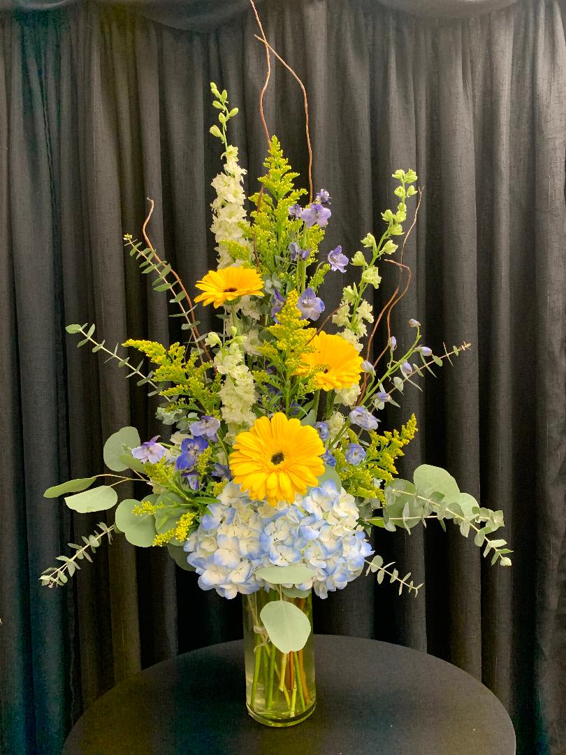 Tall bouquet of yellow gerberas, blue hydrangea, and purple flowers in a glass vase