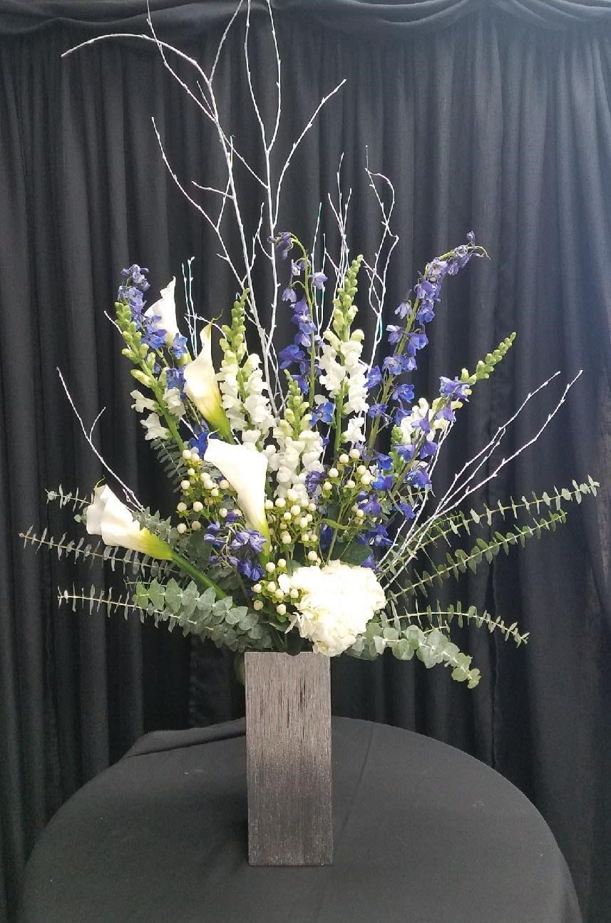 Tall floral arrangement with white calla lilies and blue-purple spikes in a wooden vase