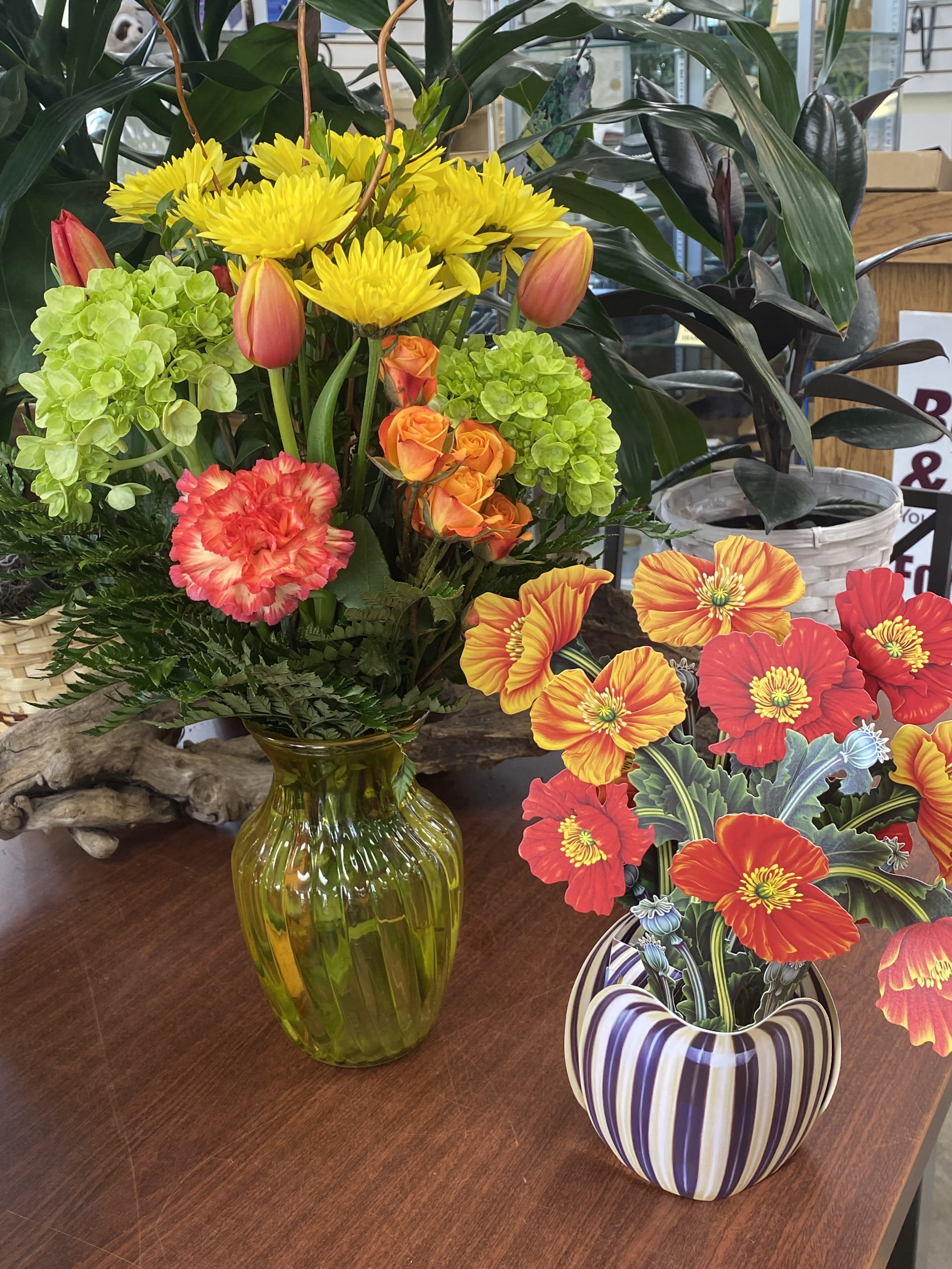 Bright mixed flower arrangement in a green glass vase beside a striped ceramic pot