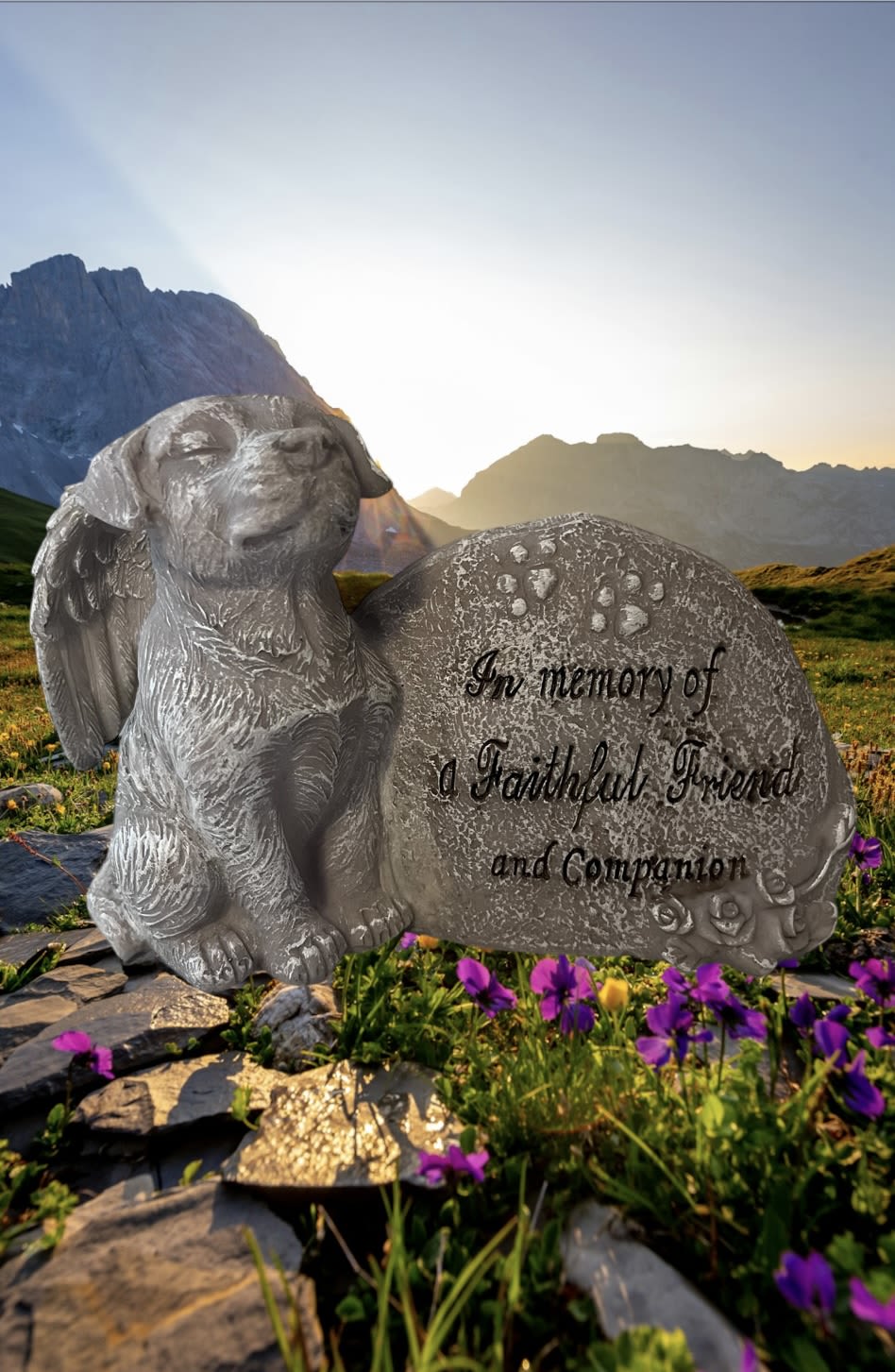 A memorial dog statue beside an engraved stone in a mountain meadow.