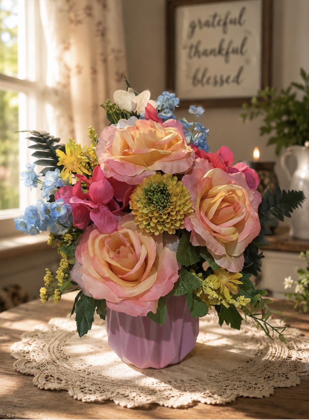 (Silk) Pink Sunrise  - Soft pink-and-peach roses take center stage in this cheerful birthday bouquet, arranged around a round yellow-green focal bloom that brightens the composition. This celebratory everyday gift - perfect for birthdays, thank-you gestures, or spring occasions - also includes blue delphinium and small yellow accents woven through for a light, feminine palette that feels joyful and fresh for the person you're thinking of.  The full, rounded arrangement is presented in a ribbed pink ceramic vase that gives it a polished tabletop presence ideal for dining tables or office desks. Alongside the roses and delphinium, delicate pink hydrangeas and mixed daisies add texture and airiness; the mix of colors-pink, peach, blue, and yellow-creates a soft, uplifting display. Sized to be a noticeable centerpiece without overwhelming a small space, this arrangement arrives ready to enjoy and makes someone feel genuinely celebrated.