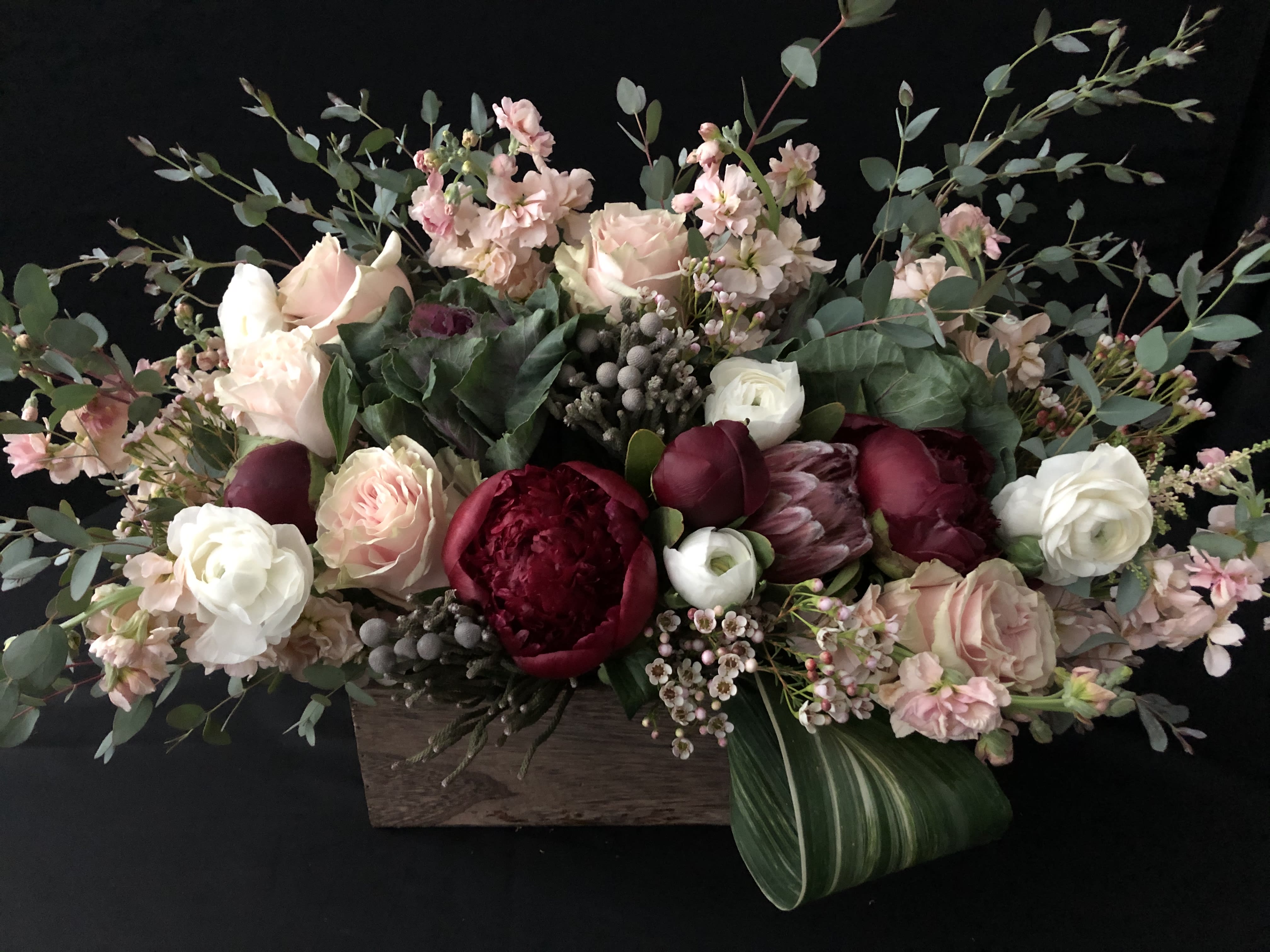 Low wooden box arrangement with blush roses, white ranunculus, and deep red blooms in a lush, elongated design