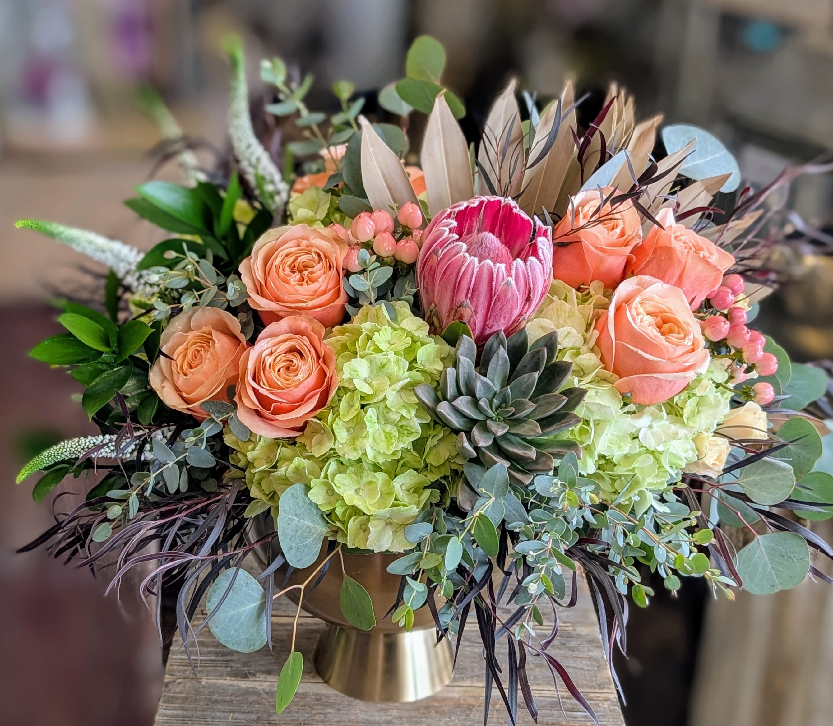 Claudia - A intriguing mix of roses, protea, hydrangea, a succulent, veronica and mixed greenery in a gold footed compote.