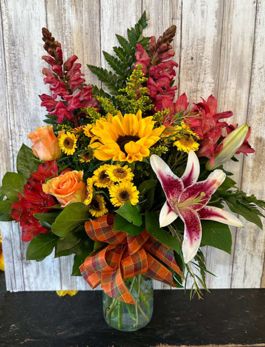 Fall Mason Jar One Sided (Substitutions Will Be Necessary) - Send this Mason Jar filled with Fall Flowers to brighten their day! Fall leaves, roses, mums, sunflowers and lilies are just a few of the fall blooms in this arrangement!  Depending on Availability, substitutions may be made. Arrangement is one sided.  The deluxe version will be made all around and will differ from the photo. 