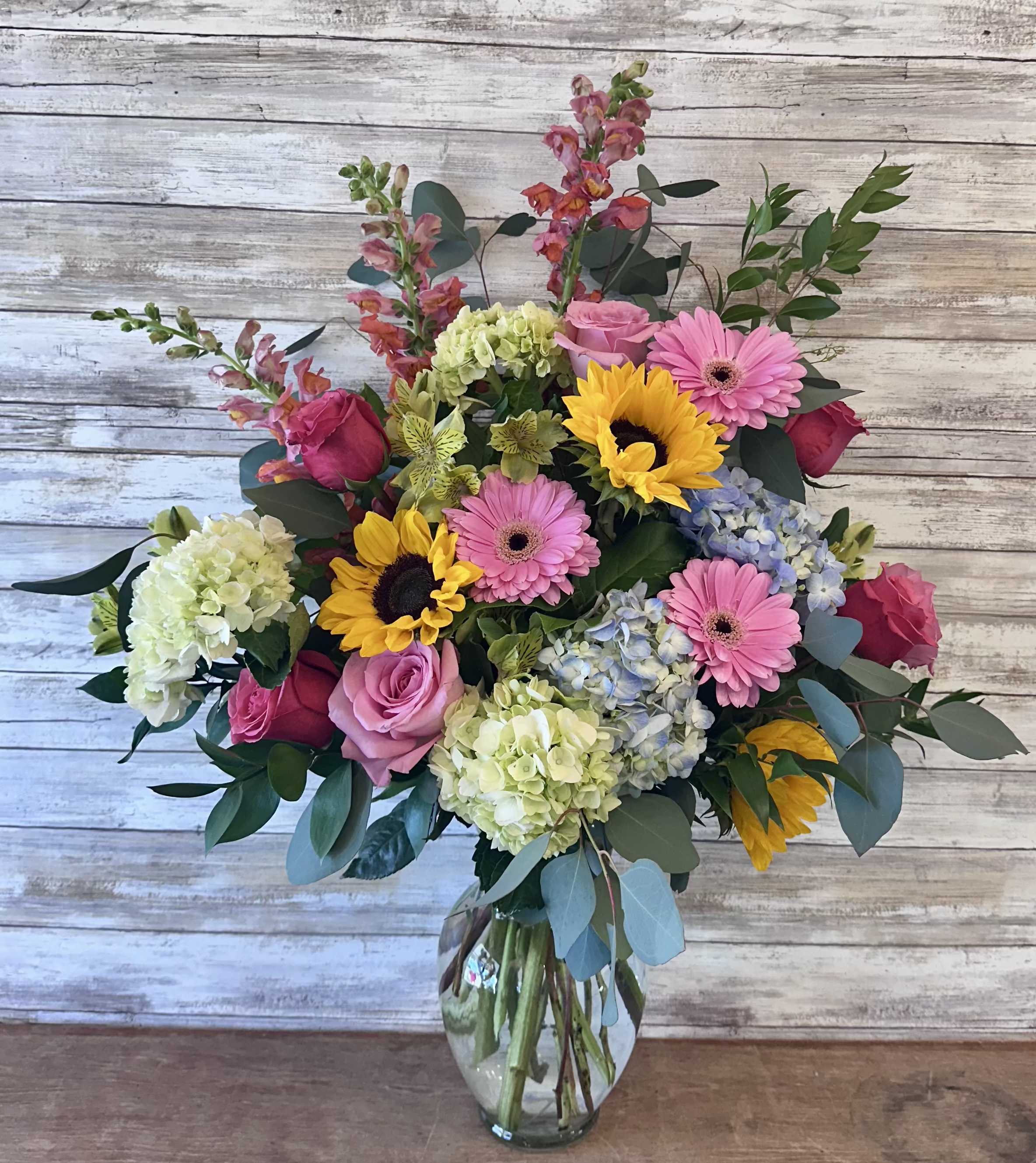 Mixed bouquet of pink roses, gerbera daisies, sunflowers, and hydrangeas in a glass vase