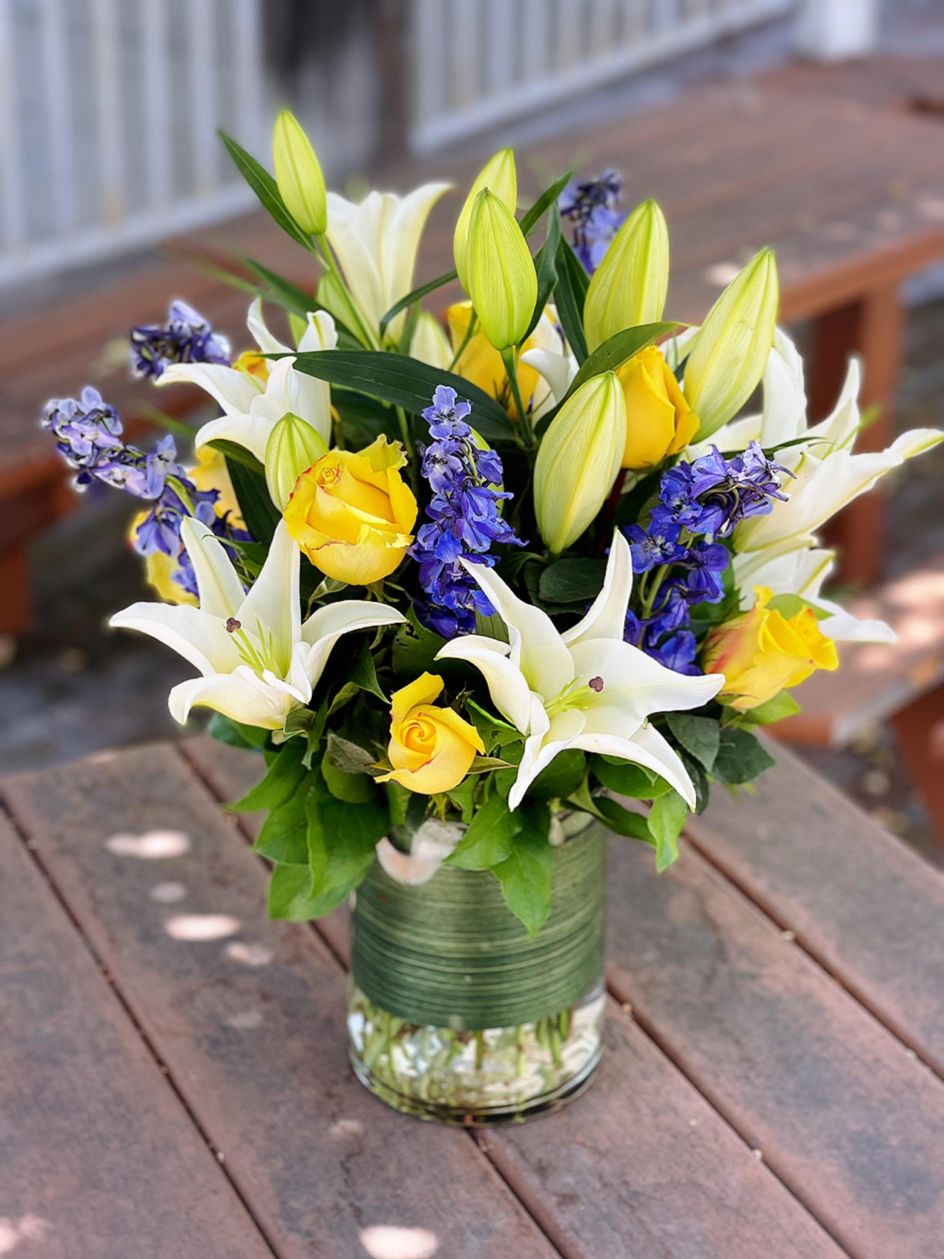 Just Bloom - - 3 Stems White Lilies - 6 Yellow Roses - 3 Blue Delphinium - 5 x 8 Inches Cylinder Vase