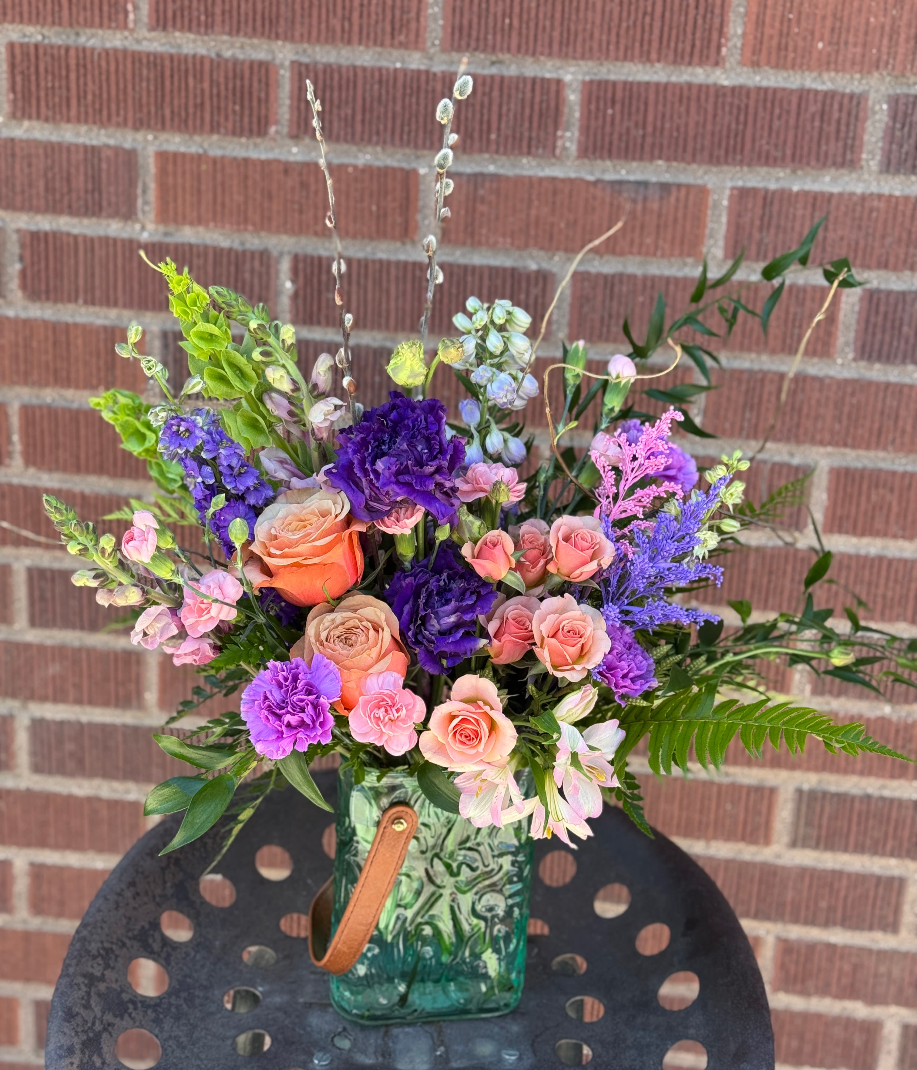 Sound of Spring - Peach and blush roses meet vivid purple carnations in a lively, garden-style bouquet designed to brighten any spring celebration or birthday - a mixed-color flower arrangement that reads cheerful and expressive. Tall spikes of purple stock and green-accent blooms give height and movement so the person you're sending it to will feel noticed and celebrated; this is the kind of arrangement that says "thank you" or "I love you" with warmth and personality.  Loose, airy branching stems and varied textures create a fresh, hand-gathered look. The bouquet pairs soft pink and peach roses with purple carnations and stock, arranged for contrast and flow, and is presented in a green glass vase finished with a leather handle for a rustic, handcrafted touch. Sized to sit beautifully on a table or entry console, it brings color and scent into a room while feeling effortlessly springlike - simple care: fresh water and a cool spot will keep the stems looking their best for days.  VASE COLOR VARIES! May be teal, purple or pink. :)