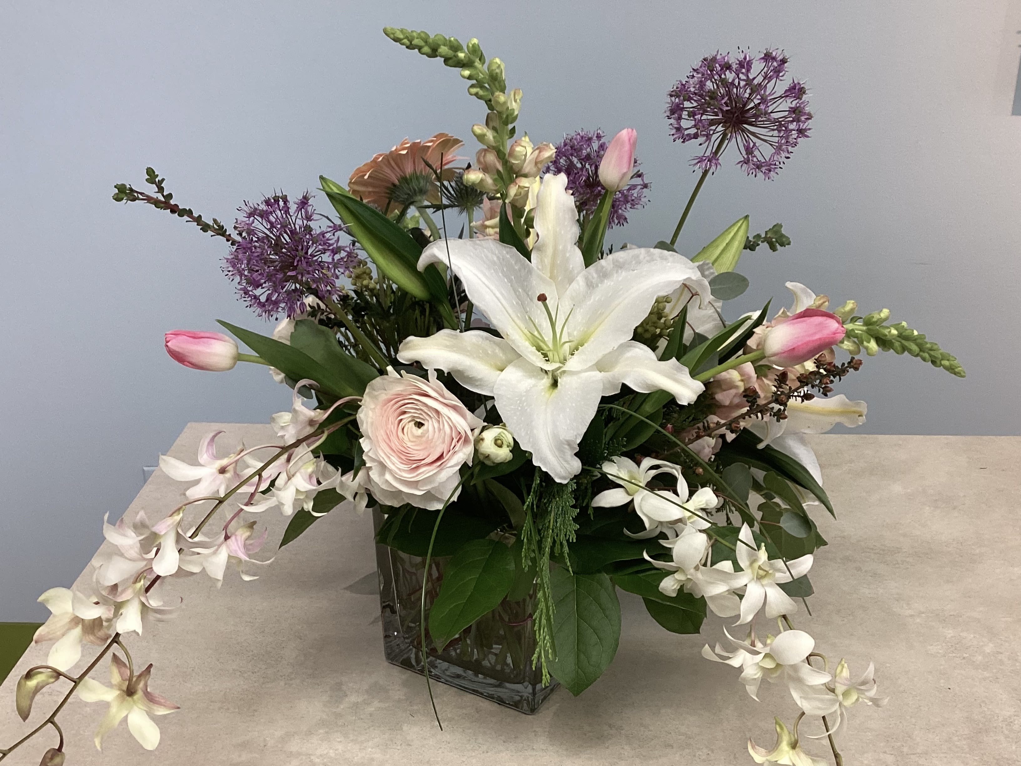 Endless Waltz - A show-stopping mix of soft pinks, crisp whites, and lifting purples, this flower arrangement is a romantic everyday gift perfect for a birthday, anniversary, or a thoughtful "thinking-of-you" surprise. The design reads like a garden bouquet-airy, outward-spreading stems and arching lines that let each bloom breathe-so the person you're celebrating will feel gently adored rather than overwhelmed.  The bouquet features large gerbera daisies, focal lilies, tulips, pale pink roses and ranunculus, purple allium-like accents, and cascading white orchid sprays arranged in a clear square glass vase. The combination of round, ruffled blooms and tall, sculptural stems creates pleasing texture and movement; soft blush and white tones are brightened by the deeper purple accents for a fresh, refined look. Dimensions suit a tabletop or entry console and the clear glass keeps the presentation modern and light.  Designed to make someone feel truly seen, this arrangement works beautifully for moms, partners, close friends, or anyone who loves a soft, garden-inspired palette. Change the water every few days, recut stems at an angle, and keep away from direct heat to extend bloom life-easy care so they enjoy the flowers longer.