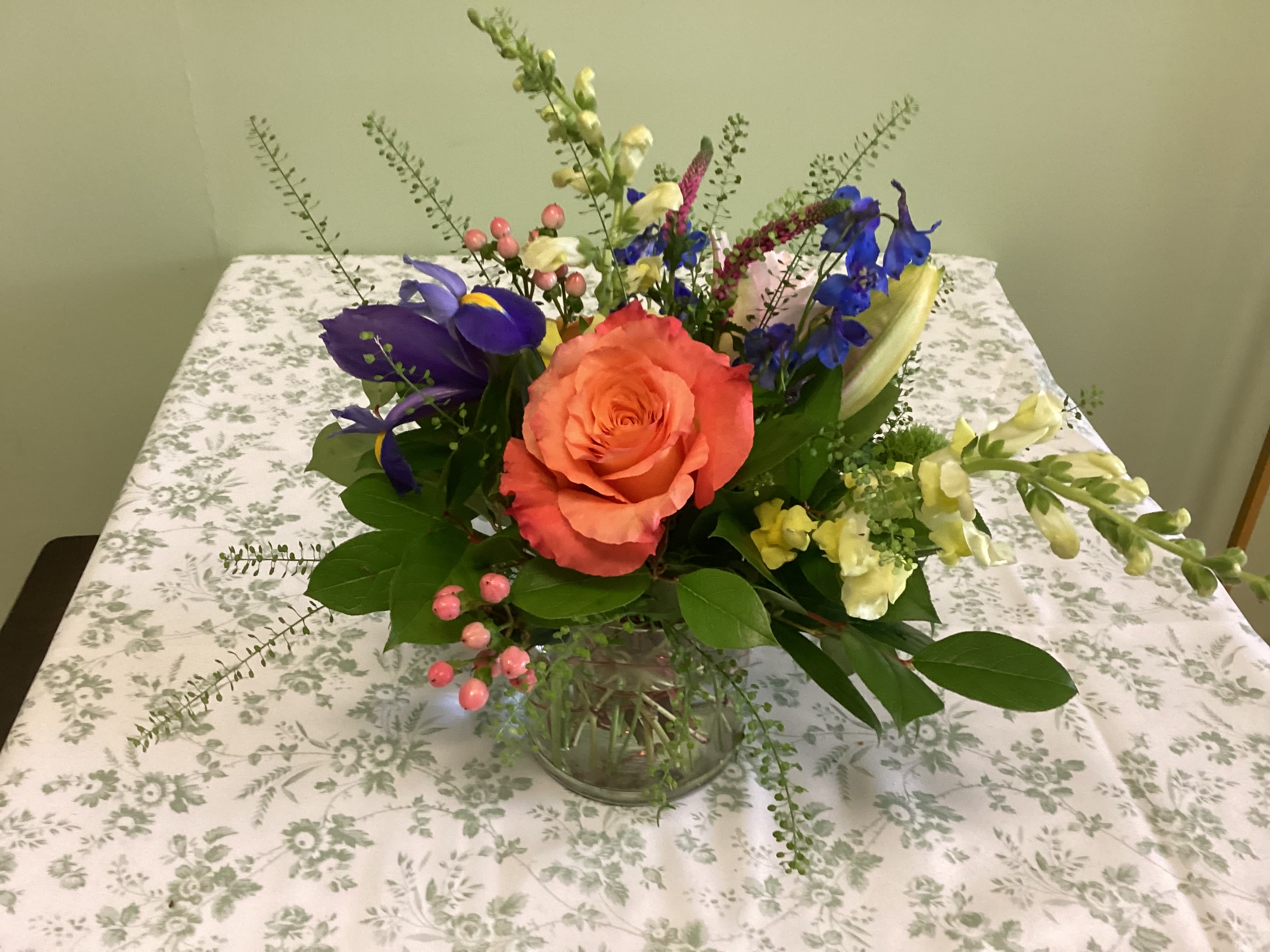 Rocky Mountain High - A cheerful, garden-style flower arrangement that makes the recipient feel genuinely seen - perfect as a birthday bouquet, thank-you gift, or an everyday just-because surprise. This flower arrangement centers on large roses and seasonal focal blooms to create a bright mixed-color display that reads joyful and fresh; mention it as a birthday bouquet or spring celebration to match searching customers' intent.  The mix includes roses in peach and orange tones, vivid blue irises and delphinium, pink lilies, snapdragons and tall accent spikes, plus airy filler stems and small berry-like accents for texture. Presented in a clear glass vase, the design is loose and airy with tall accents rising above the main cluster and broad green leaves framing the base, creating a lively, garden-picked feel that's both full and casually elegant.  Ideal for the person you want to lift up-she'll feel celebrated and noticed when these warm pinks and oranges contrast with cool blues and purples. Sizes are arranged to fill the vase with outward-reaching stems for a natural silhouette; order now for same-week delivery and simple care: fresh water, a cool spot out of direct sun, and a trim of stems every few days to keep the bouquet vibrant.