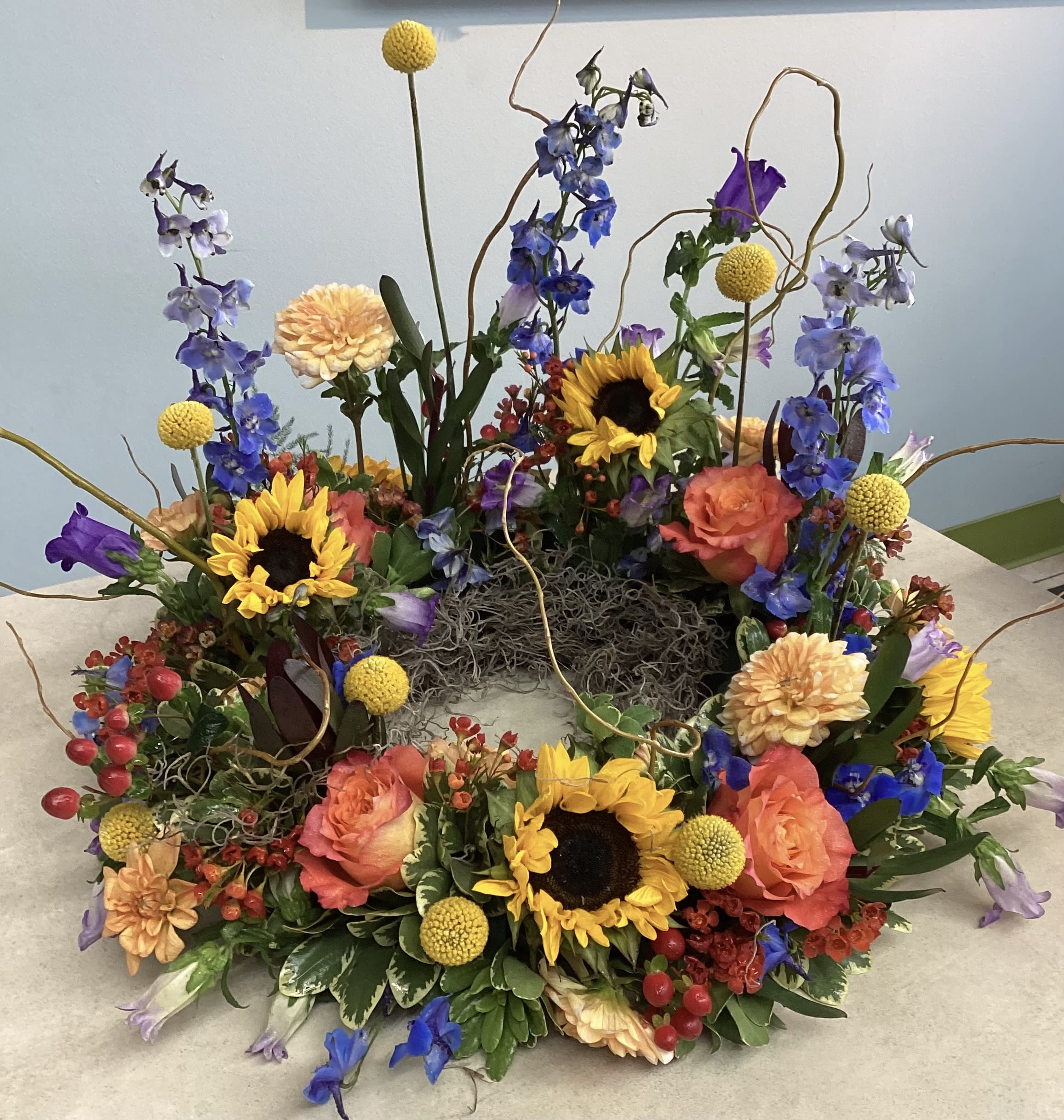 Colorado Wildflower Sympathy Urn Wreath - Bright mixed wildflowers including sunflowers, roses, dahlias and blue delphinium, arranged with moss and curly willow branches for a fresh sympathy wreath in yellow, ,orange and blue.