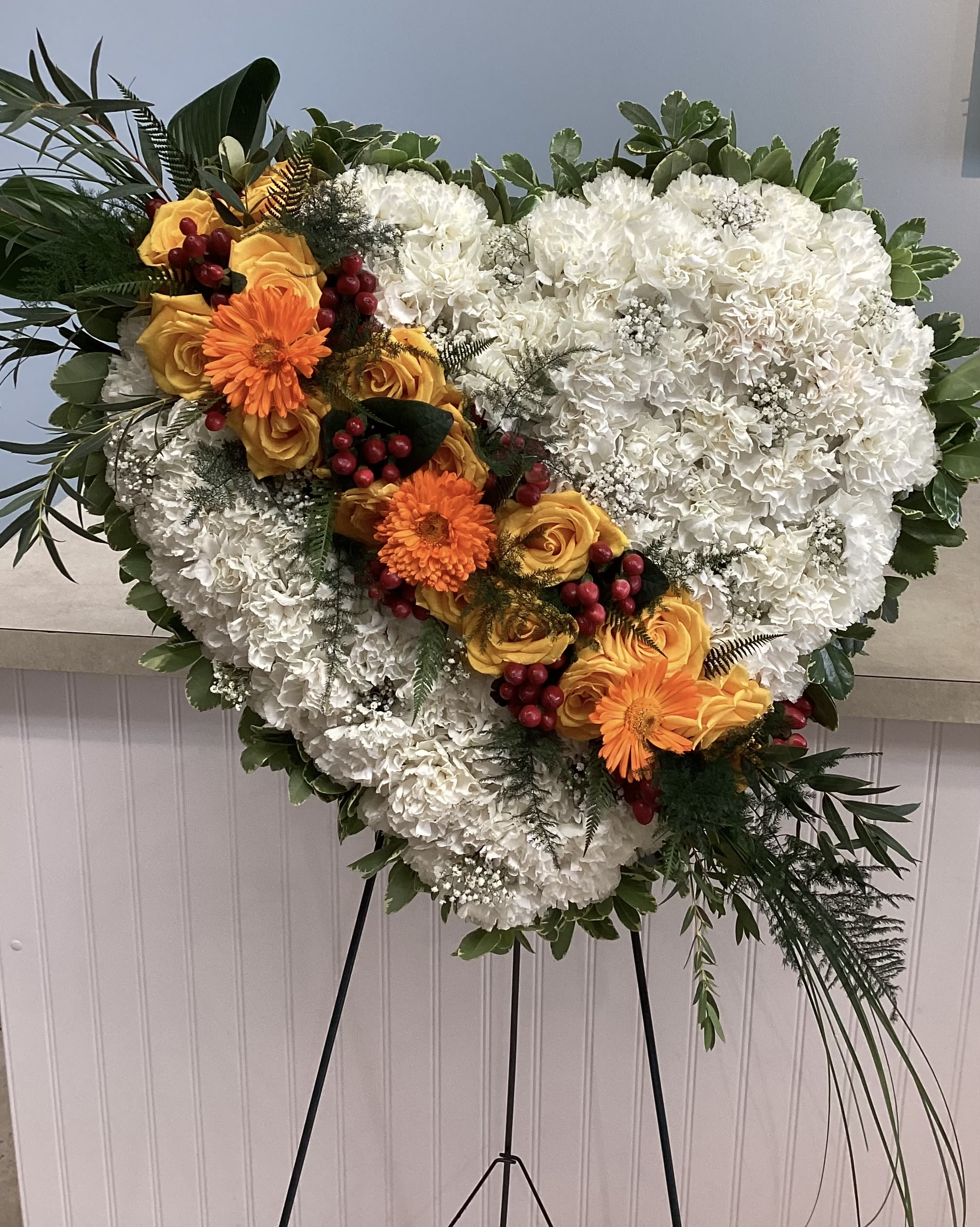 Standing Heart - White heart-shaped standing spray of carnations with a diagonal band of orange gerbera and yellow roses, red hypericum berry accents and greenery-formal tribute for a memorial service.