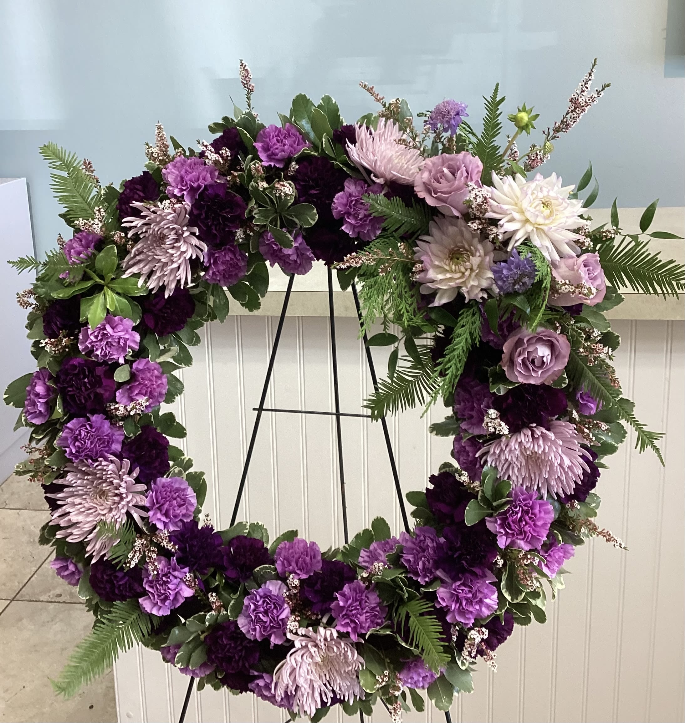 Lavender Fields Standing Wreath  - Purple, lavender and white funeral wreath of chrysanthemums, carnations and roses with greenery, densely packed and mounted on a black easel for an elegant, somber memorial tribute. 