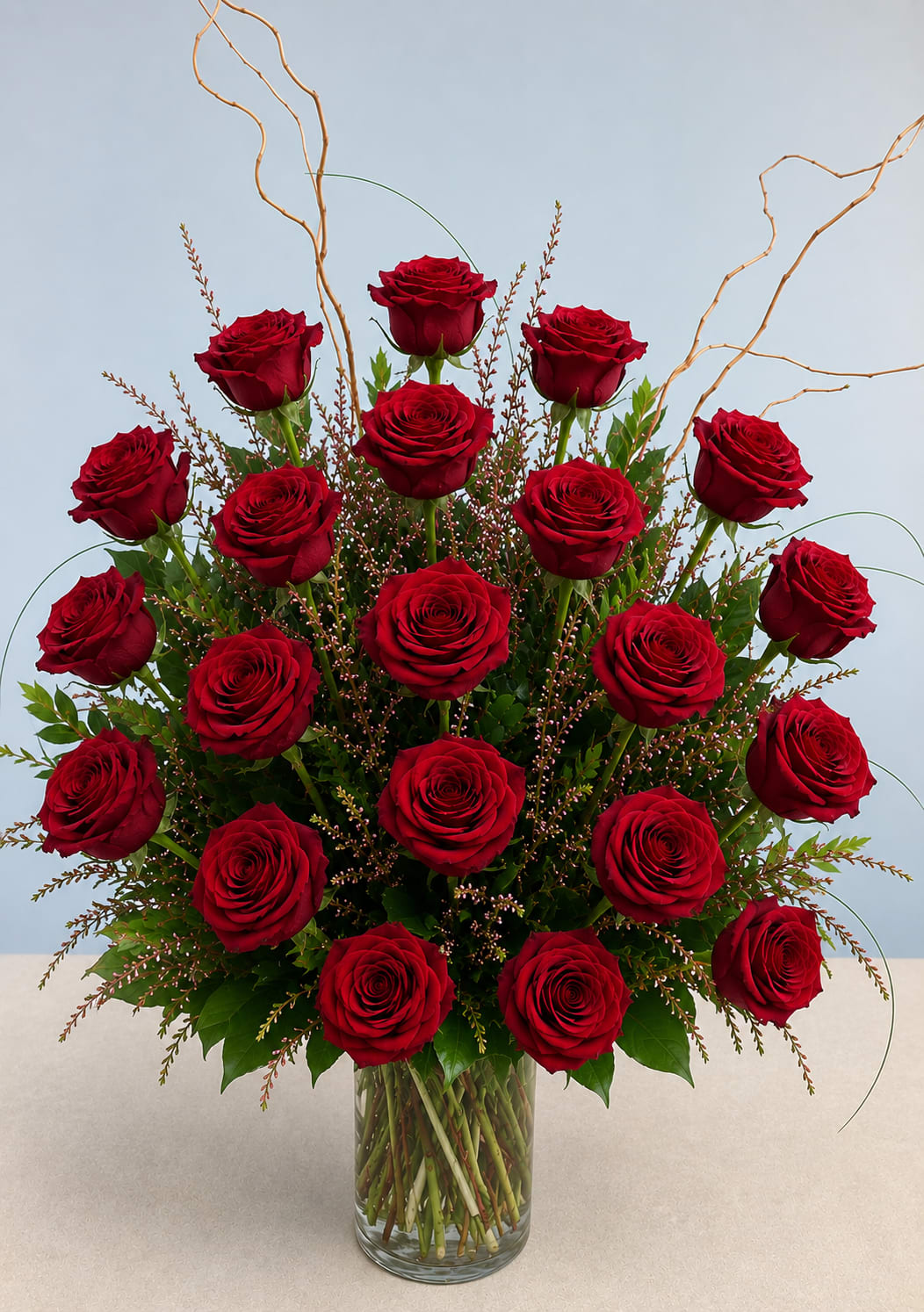 24 Red Roses - Velvety, deep red roses with textured greenery in a tall clear vase, accented by curly willow branches and filler for a classic, romantic arrangement perfect for love or celebration.