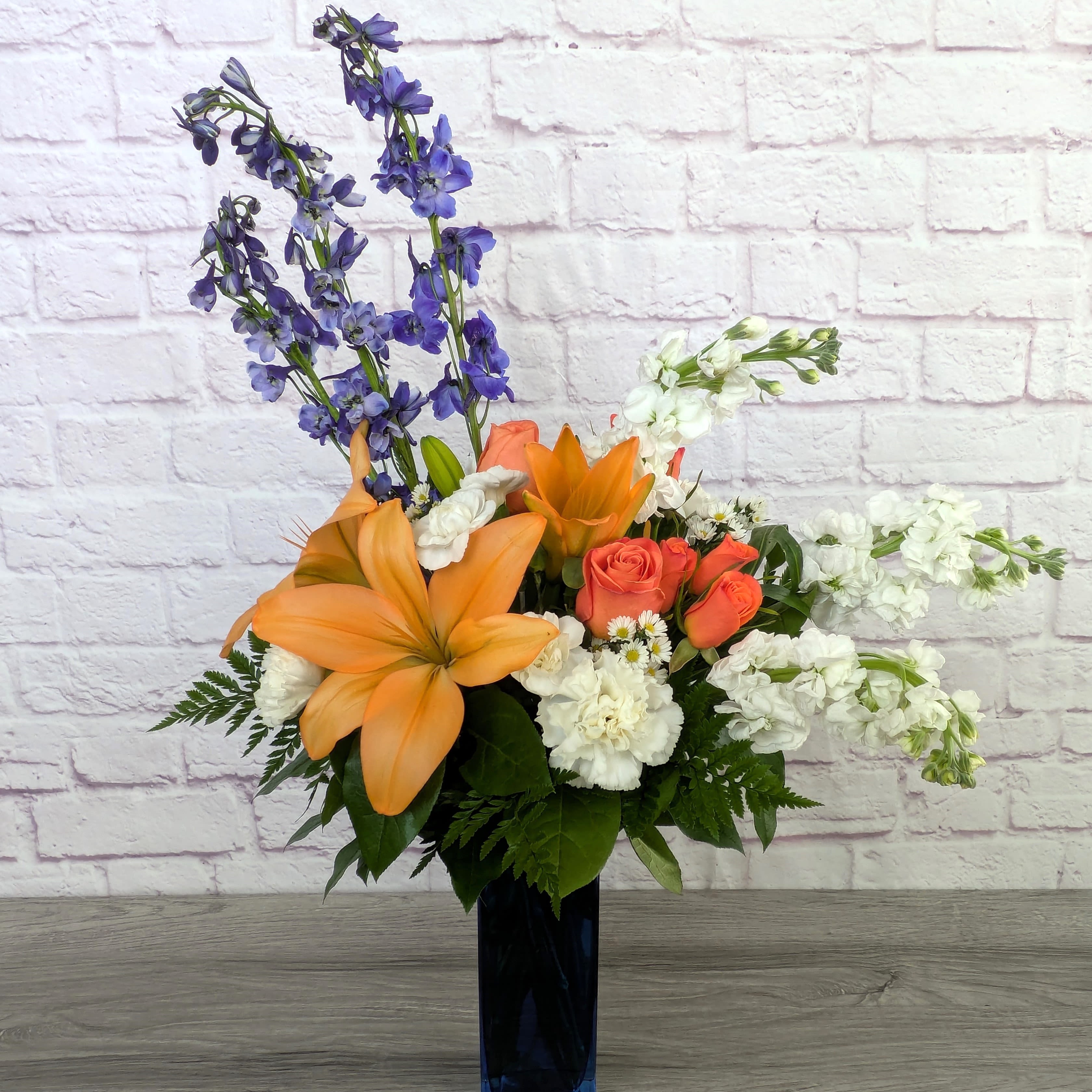 Orange lilies and roses in a tall dark vase with white and purple flowers