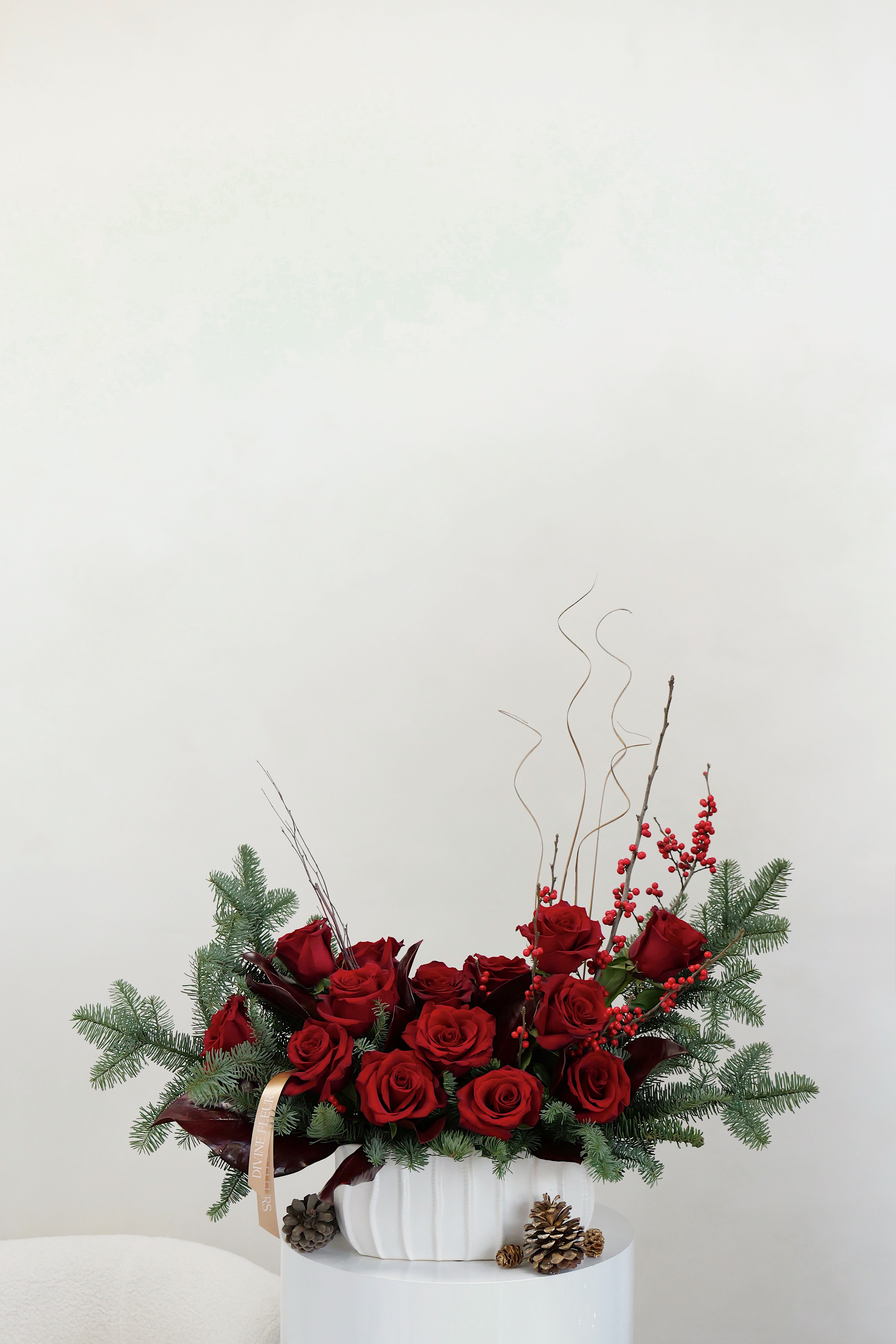 Red roses arranged in a white vase with pine branches and berries