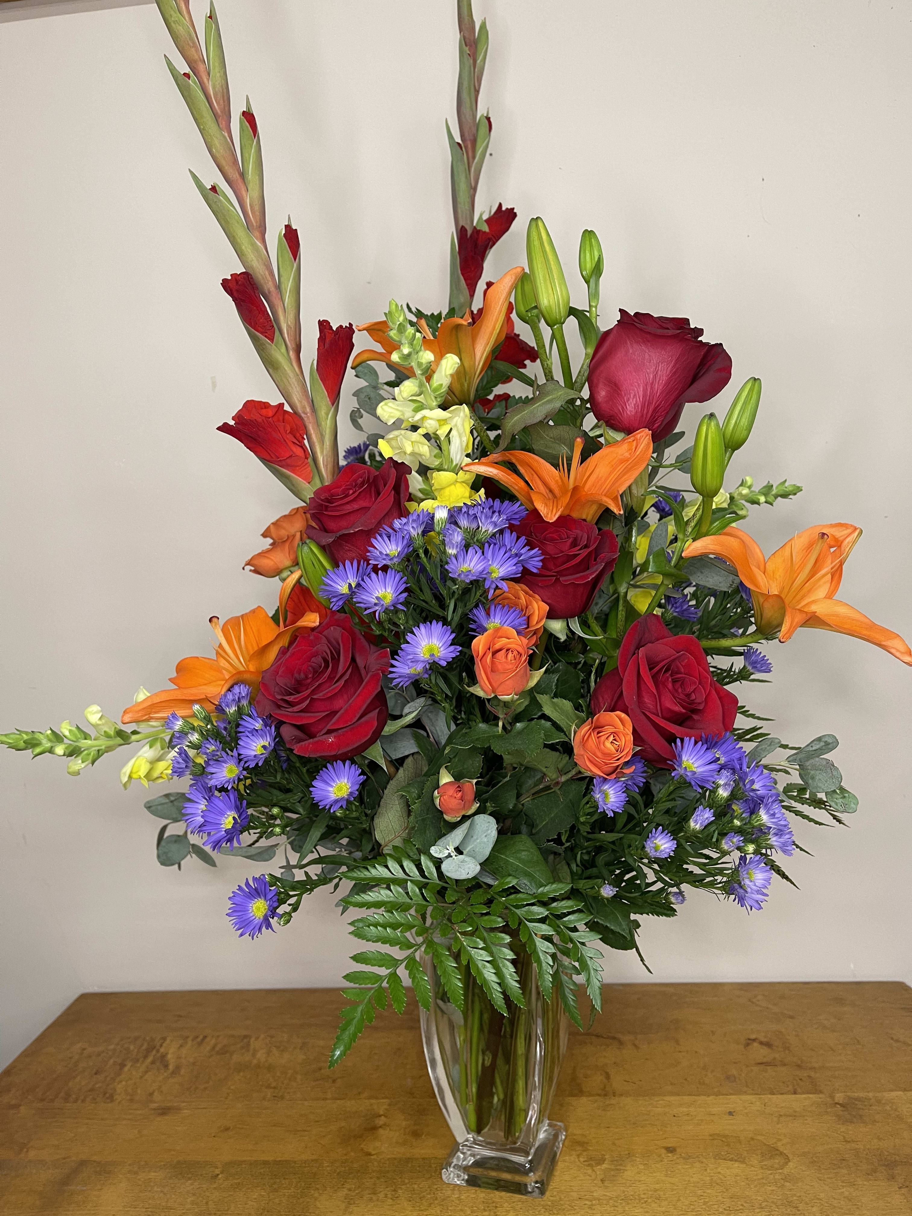 Mixed bouquet of red roses, orange lilies, and purple daisies in a clear vase