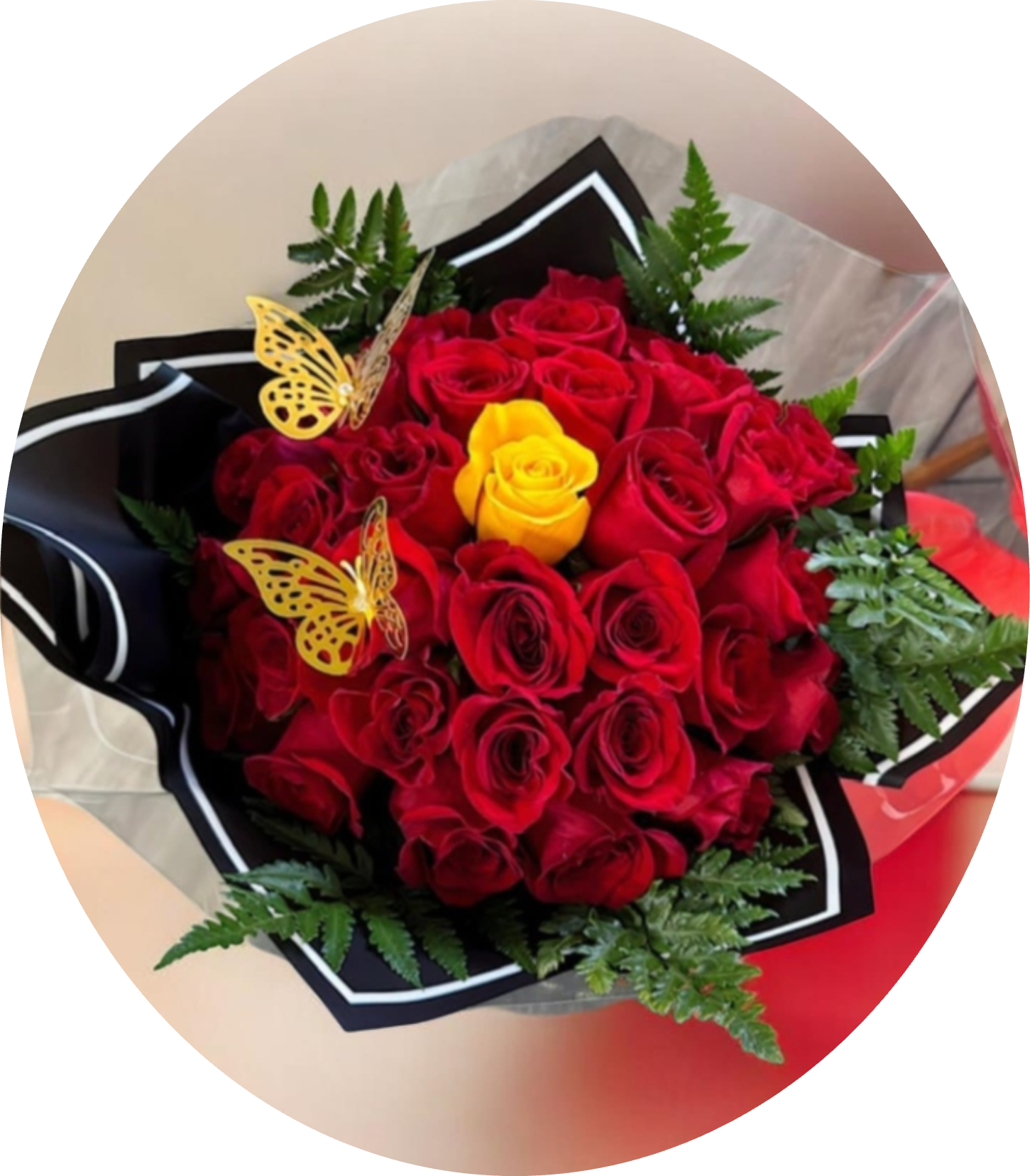 Macarena RAMO BUCHON RED ROSES - A dense cloud of red roses punctuated by a single bright yellow rose creates an immediate focal point-perfect for an anniversary bouquet or a bold Valentine's Day gift. The high-contrast black-and-white wrapping and clear cellophane give this arrangement a modern, dramatic look that reads romantic and celebratory; the person you're sending it to will feel noticed and adored the moment it's handed to them.  Framed with fern-like green foliage for softness and finished with decorative butterfly picks, this bouquet balances texture and playful detail. Wrapped in layered black paper with white trim and a clear outer plastic, the presentation stays polished from delivery to display. Sized as a compact, tightly clustered arrangement, it's an excellent choice for anniversaries, birthdays, or any occasion when you want someone to feel truly seen and celebrated.