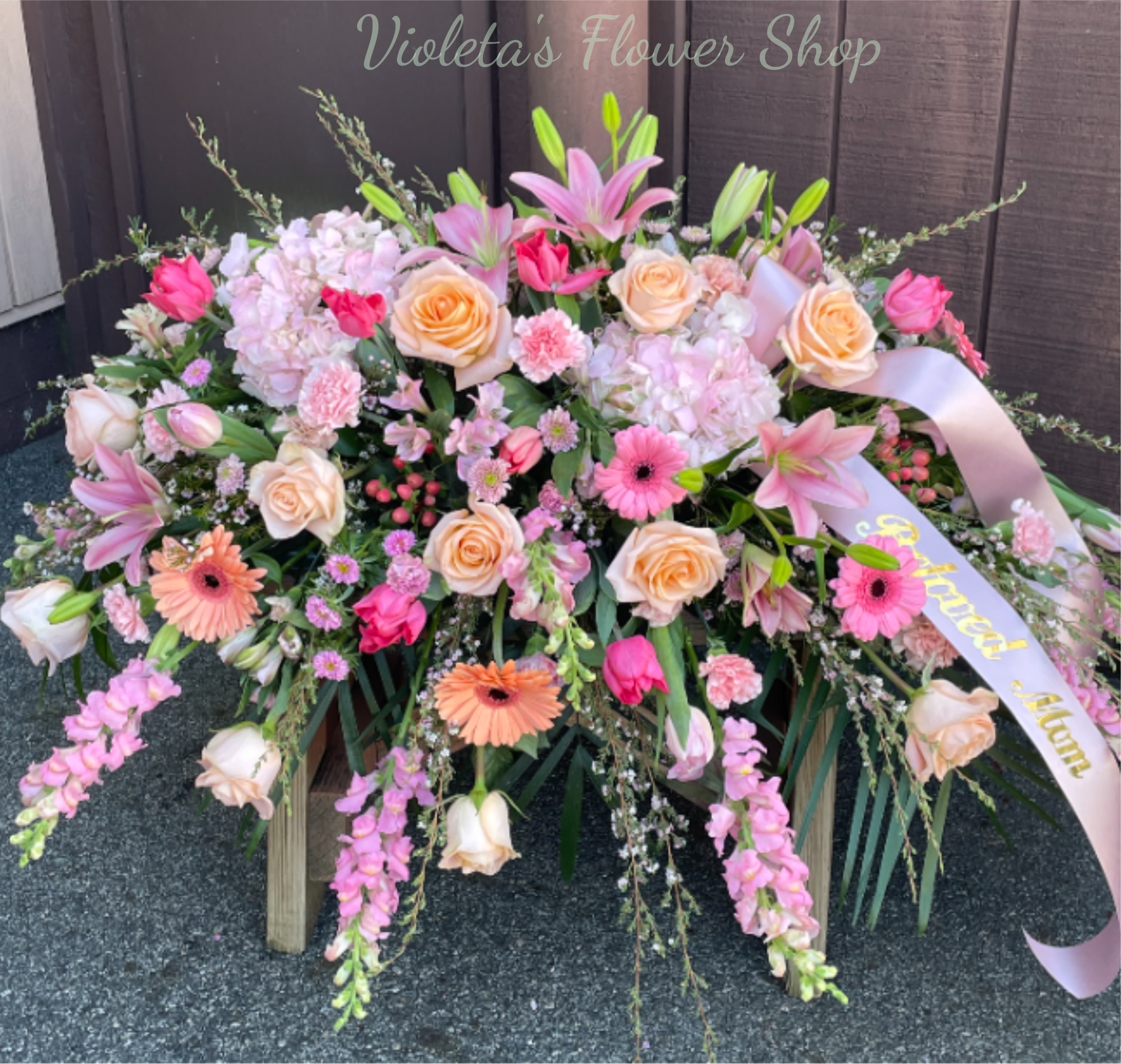 Soft Pastel Blooms Casket Spray - A generous, low-spreading casket spray in soft pinks, peaches, whites, and fresh greens creates an immediate sense of calm and tenderness - ideal as a sympathy casket spray or as a formal memorial tribute.   This abundant spray features a dense mix of roses, lilies, gerbera daisies, carnations and trailing snapdragon-like stems arranged in layered, textural waves for depth and visual movement. Finished with a pale pink satin ribbon and presented on a sturdy wooden stand, the composition sits low but wide for a dignified, photographed-ready presentation. The pastel palette of blush, peach and cream keeps the tone gentle and comforting while the green foliage adds fresh contrast.  