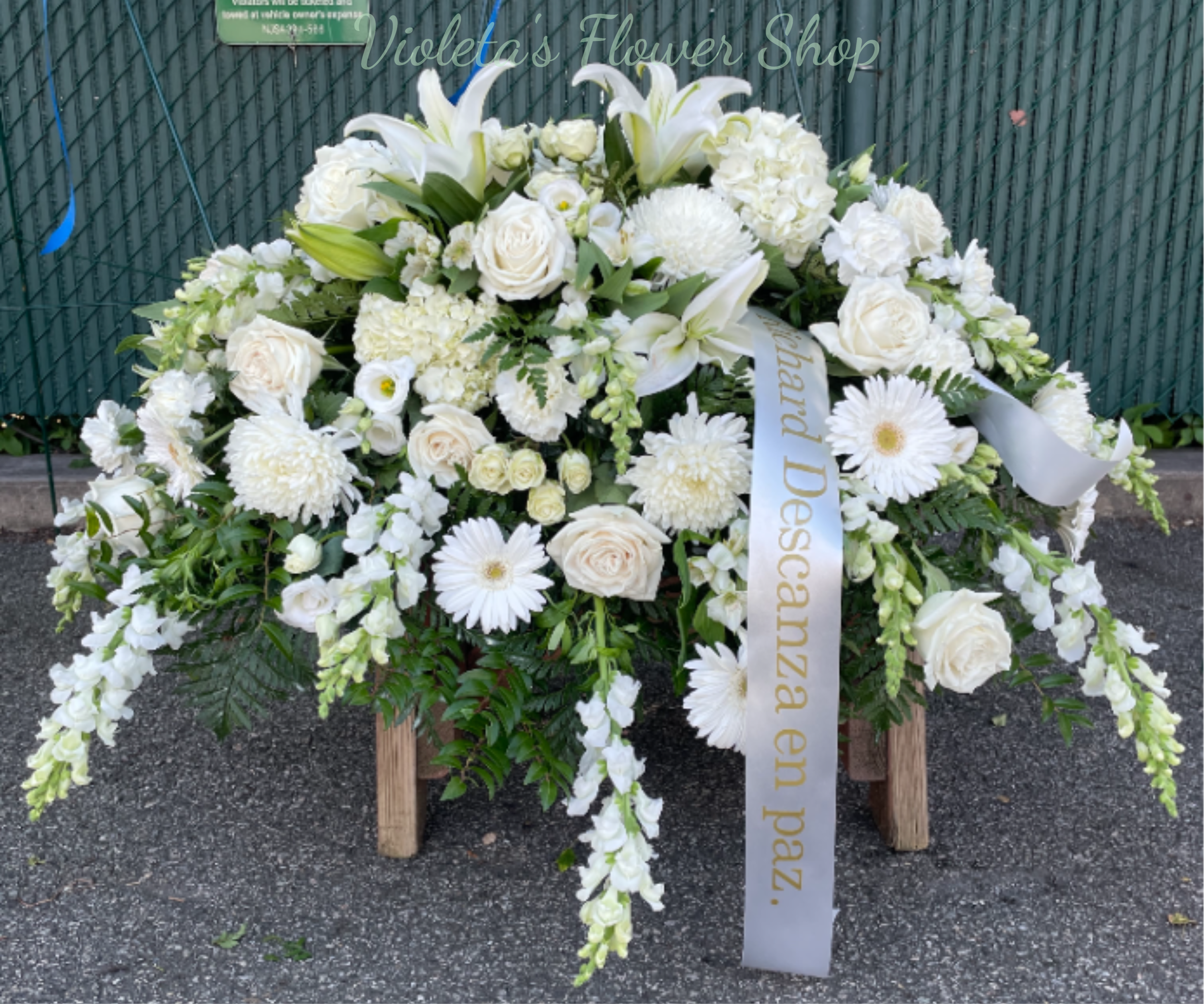Casket Spray - A cascading white casket spray that reads as serene and respectfully formal - ideal for sympathy and funeral services when you want the person you're honoring to be remembered with quiet dignity. This large spray is displayed on the casket and framed with a long mourning ribbon (white with gold lettering) to reinforce the memorial presentation; it gives the mourners a focal tribute that looks composed from every angle.  The arrangement combines white roses and lilies with daisy-like blooms, chrysanthemums, and trailing snapdragon-style spikes set into soft green foliage for layered texture and depth. The dense, cascading shape creates a full, continuous silhouette across the casket; flowers are arranged for lasting visual impact and a balanced mix of open blooms and vertical accents for contrast.  For use at visitation, service, or graveside, this spray speaks to comfort and remembrance - the kind of tribute that helps family and friends feel seen in their grief. 