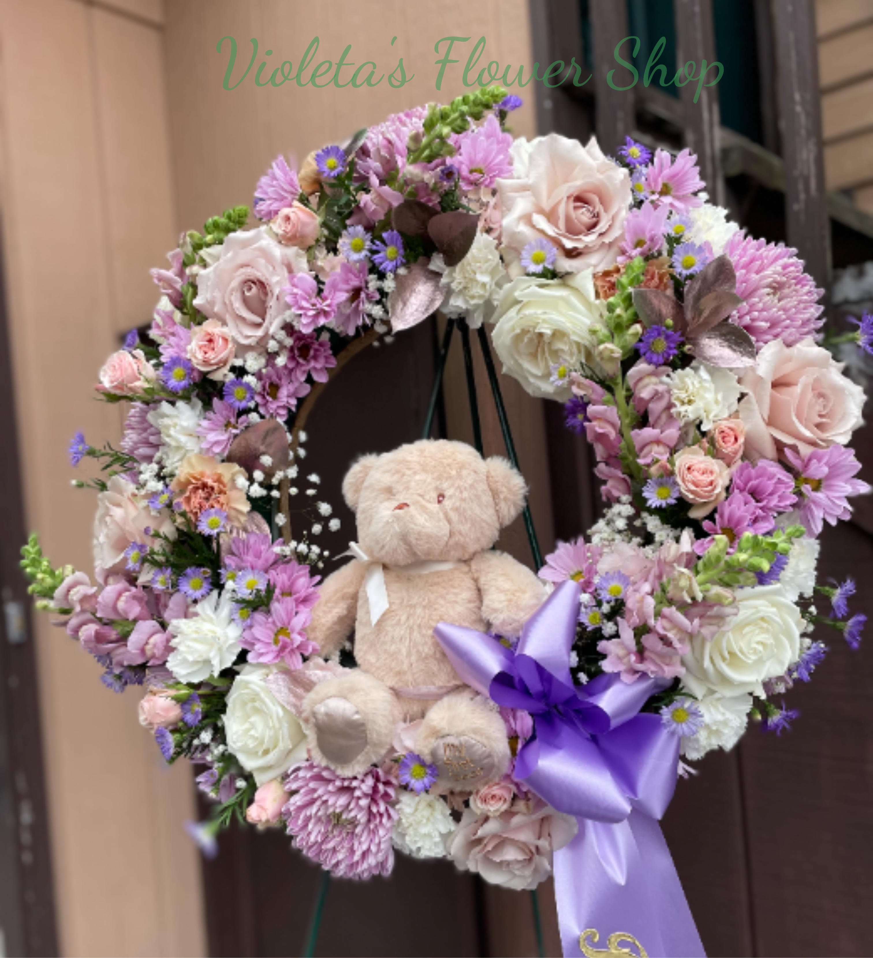 Tender Memories Wreath - A plush teddy bear nestled at the heart of a round sympathy wreath gives Tender Memories Wreath an immediately tender, comforting presence-perfect as a sympathy or funeral tribute that also reads as a gentle keepsake for a family remembering a loved one. This flower arrangement wreath pairs soft pink, white, and lavender roses, carnations, and daisy-like chrysanthemums to create a layered, delicate palette that speaks to consolation and warmth. The purple satin ribbon adds a graceful, gift-like finish that helps the piece feel personal and thoughtful.  Choose this wreath when you want the recipient to feel gently seen and comforted; it's an especially meaningful way to honor someone at a funeral or memorial while offering a keepsake the family can cherish. For ordering, specify delivery date and service details at checkout so the wreath arrives freshly arranged and ready to display on the included easel.