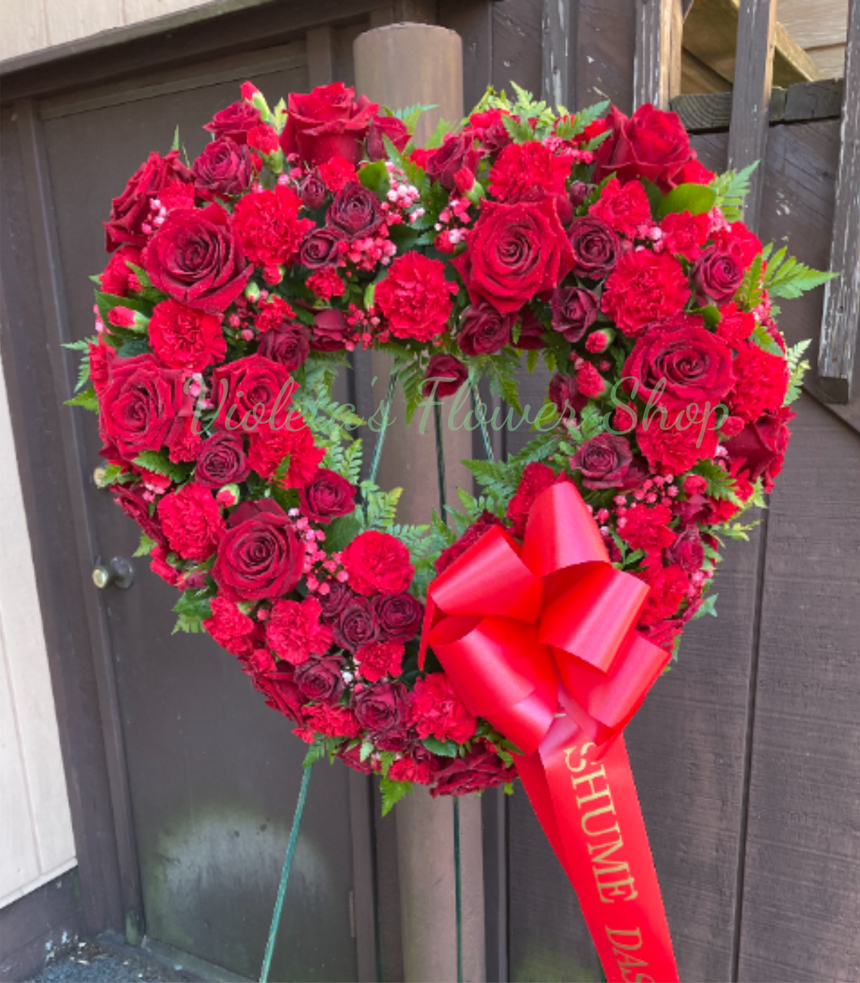 Until We Meet Again Open Heart - A lush heart-shaped sympathy arrangement that speaks for you when words fall short - perfect as a memorial tribute or a love-themed remembrance. The Until We Meet Again Open Heart arranges red roses and red carnations into a densely packed, ceremonial heart that delivers a bold, emotional statement for funerals, memorial services, or an anniversary in memory of someone dear.  The wreath is layered with spray roses and small red filler blooms woven with fern-like green foliage for contrast and depth, creating a full, romantic look from every angle. It's finished on a wire frame with a glossy red ribbon bow, a printed hanging ribbon sash, and is displayed on a wire wreath stand to present respectfully at services or at home.  