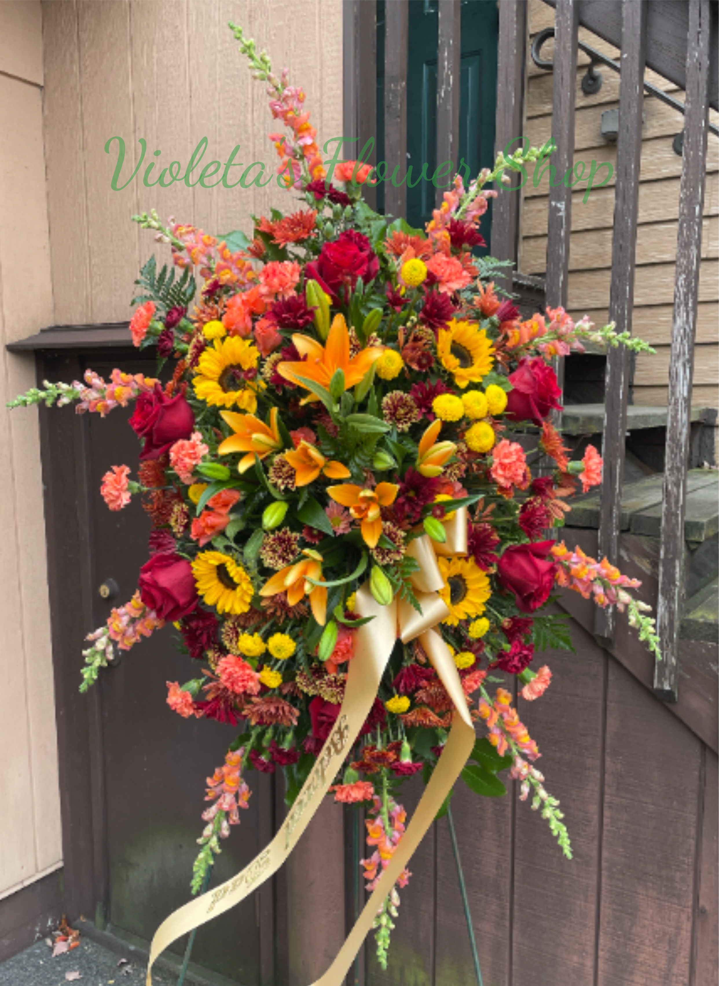 A Thousand Sunsets - A bold cascade of sunflowers, lilies, roses and warm-toned blooms makes this standing spray a striking sympathy arrangement for a memorial service or celebration of life. The large floral easel display-tied with a gold ribbon-creates an immediate, ceremonial presence that honors a loved one with bright, uplifting color instead of somber tones. Designed to stand on an easel, this piece reads as a prominent tribute that will make the person you're remembering feel celebrated and vividly remembered. Measure and placement are important for ceremony spaces-please indicate if you need help with delivery timing or easel setup when you place your order.
