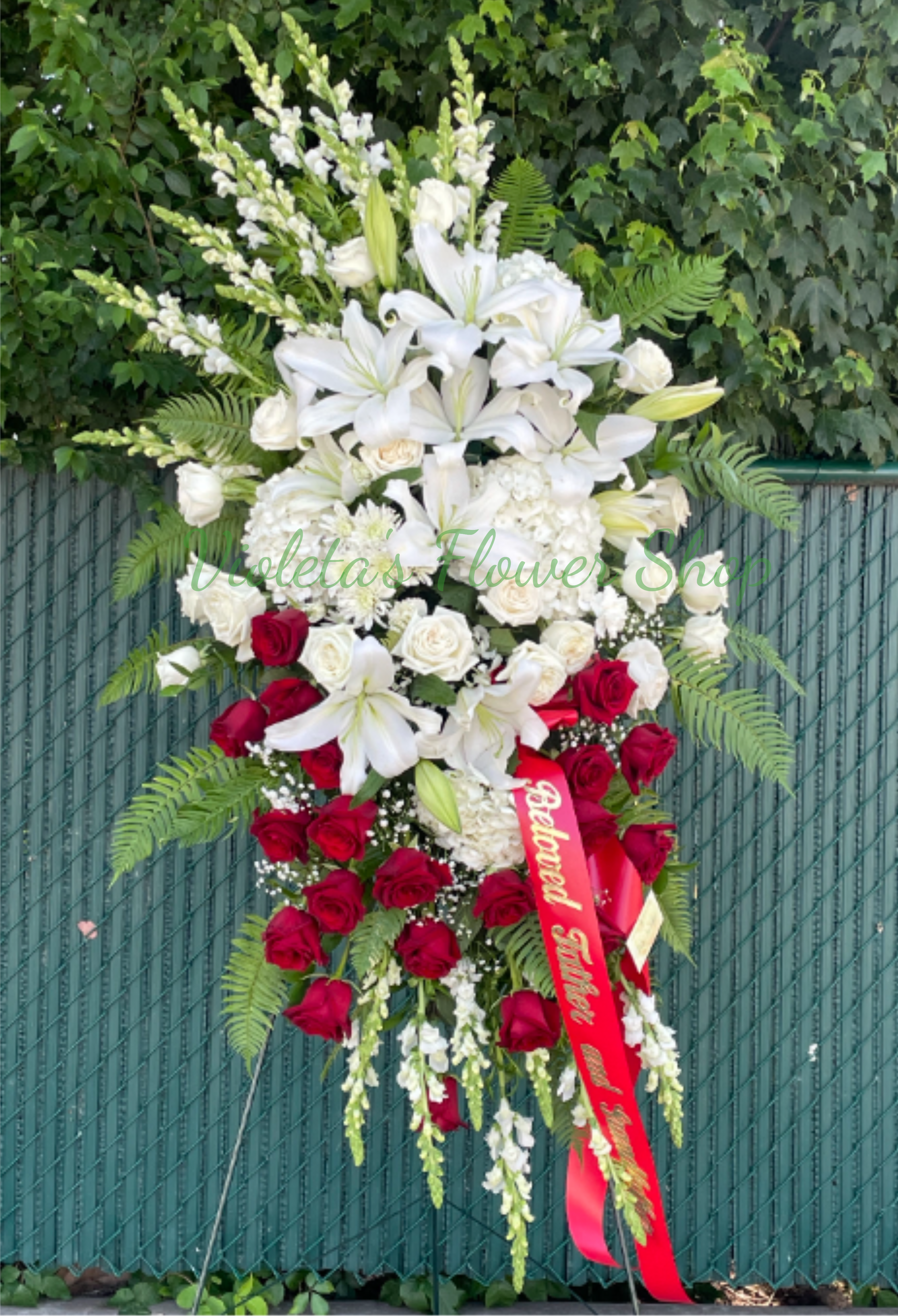 Eternal Peace Standing Spray - The arrangement pairs white lilies, white roses, hydrangea clusters, snapdragons, and chrysanthemums with rich red roses and lush green fern-like foliage for height and movement. It's mounted on a standing easel and finished with a red ribbon bearing gold lettering for a traditional ceremonial touch. The layered, tapered composition creates visual balance from top to bottom while the mix of textures-soft hydrangea clusters, sculpted lilies, and velvety roses-reads clearly from a distance.  Ideal for funeral services, memorials, or graveside ceremonies, this standing spray communicates sympathy, honor, and solemn beauty on behalf of family or friends. Available to order for delivery to funeral homes and service locations; please let us know any ribbon wording and placement preferences when you place the order.