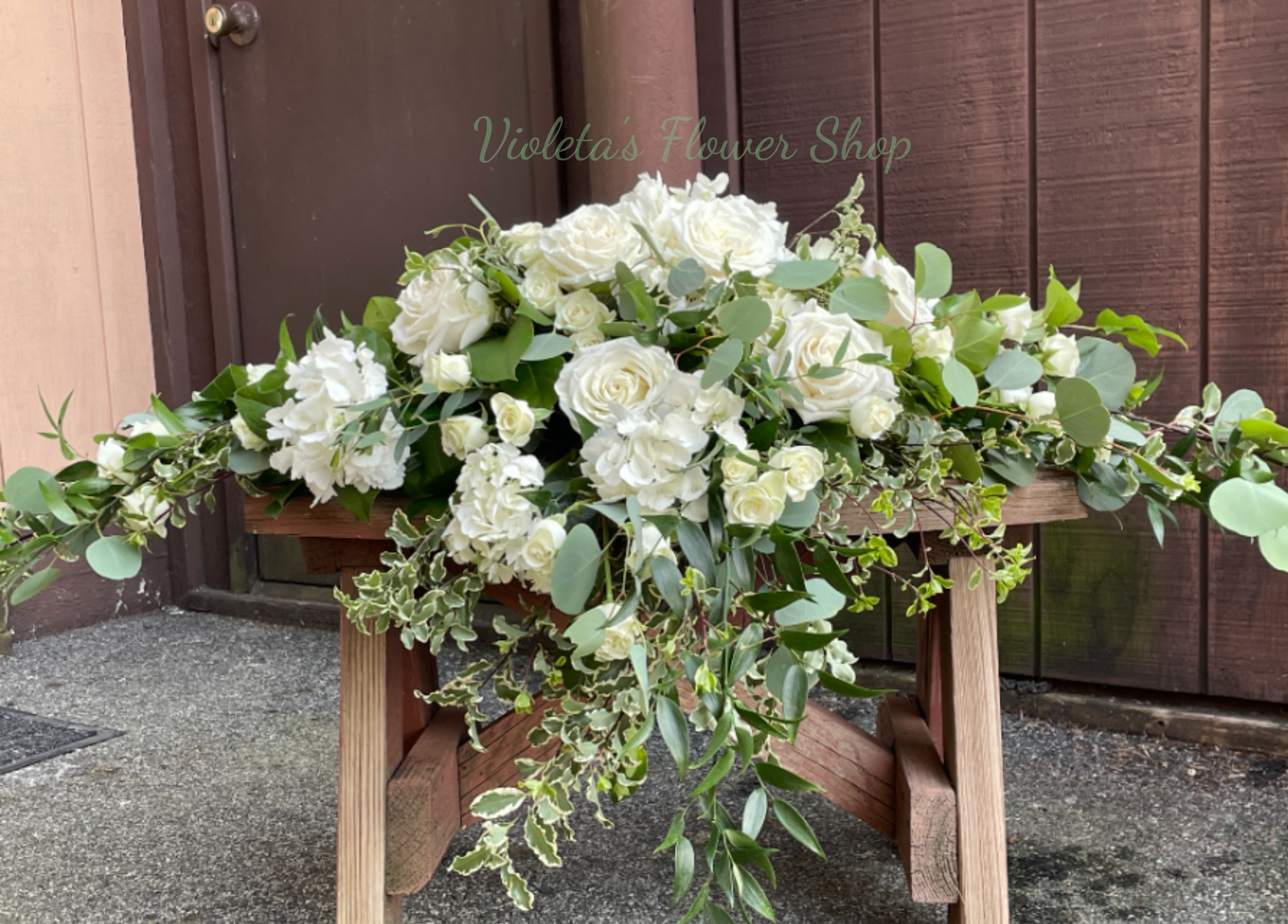 Field of Blooms Casket Spray - A broad, garden-style casket spray that unfolds like a Field of Blooms-soft white roses and delicate white filler flowers create a calm, elegant display perfect for sympathy or a formal memorial. This funeral casket spray presents a serene all-white palette that reads respectful and timeless, the kind of arrangement that helps the person you're honoring feel remembered and seen.  Low and sprawling in shape, the design is densely packed with white roses at its heart and airy white fillers that give soft texture; trailing greenery extends beyond the main body for a lush, natural silhouette. Photographed on a wooden bench to show scale, the horizontal spread emphasizes abundant fullness and a peaceful, garden-like presence suitable for viewing on a casket or as a memorial centerpiece. 