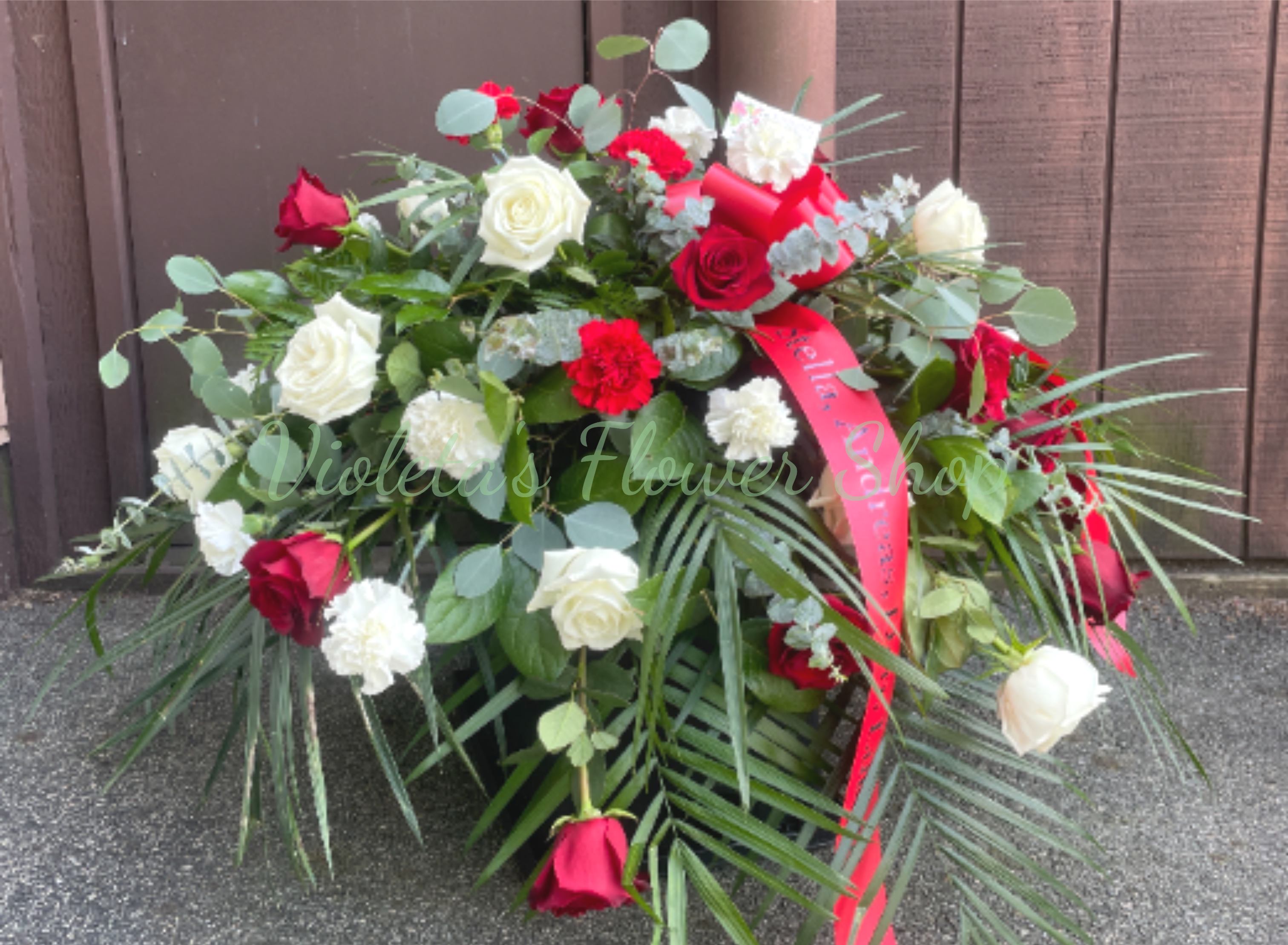 Casket Spray Roses and Carnations - A lush, garden-style casket spray built from abundant red and white roses and matching carnations sets a formal, ceremonial tone the moment it's placed. This casket spray for sympathy or funeral service (also appropriate for memorials and anniversaries) combines the classic romance of red roses with the purity of white blooms to create a powerful, respectful tribute that will make the person you're honoring feel deeply remembered.  The design is large, low, and full - layered with broad palm fronds and eucalyptus-like foliage that cascade outward for a graceful, elongated silhouette. Red and white roses mingle with red- and white-striped carnations across and a long red printed ribbon drapes the front to emphasize the ceremonial presentation. Colors: red, white, and green foliage; flowers: roses and carnations. 