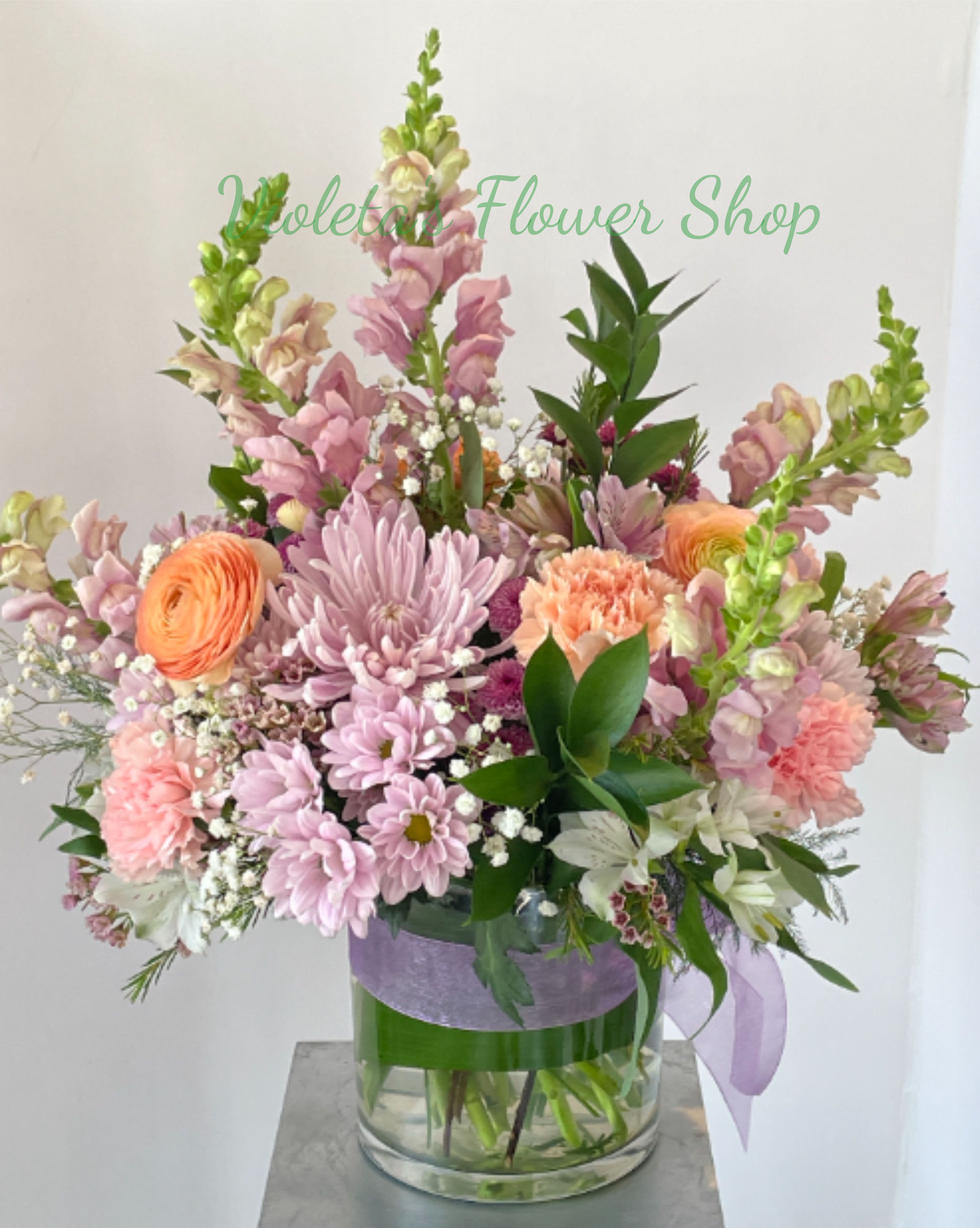 Lavender and Peach Arrangement - A full, rounded mix of chrysanthemums, carnations, ranunculus, alstroemeria and small filler blooms is arranged in a clear glass vase, with the tall snapdragons rising above a dense, textured cluster below for visual interest. The composition is finished with a wide purple ribbon and a lavender ribbon tied around the vase, reinforcing the soft color story and giving a polished, gift-ready look.  This bouquet is sized to sit beautifully on a dining table, entry console, or bedside-perfect for brightening a special day or offering comfort. Standard care: change vase water every 2-3 days, recut stems if possible, and keep away from direct heat to prolong the fresh, delicate blooms.