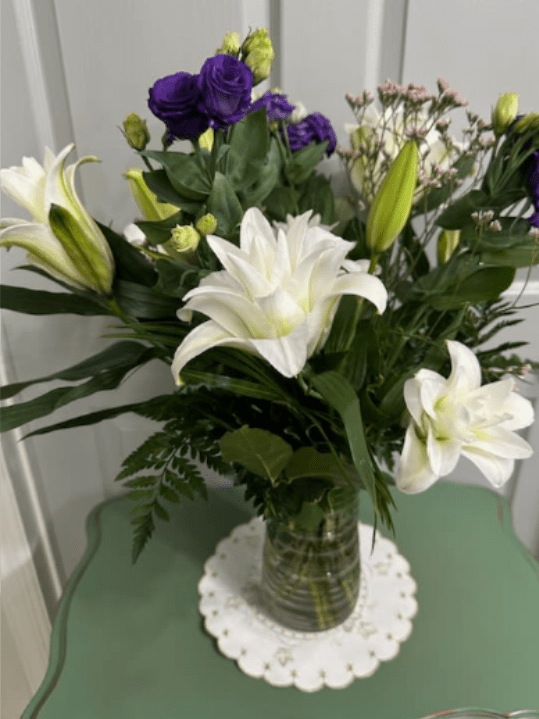 Double Bloom Lilies  Arrangement (NEW ITEM) - Large white lilies-some brushed with soft pink and green-meet clusters of deep purple lisianthus for a striking, elegant birthday bouquet or sympathy arrangement that reads both formal and tender. The upright composition places the lilies as luminous focal points while round purple accents and airy filler blooms create contrast and movement, making the recipient feel seen and comforted whether you're marking an anniversary, saying thank you, or sending get-well wishes.  Arranged in a clear or tall green-tinted glass vase with fern-like foliage and delicate white spray blooms on slender branches, this bouquet is abundant without feeling heavy. The mix of smooth lily petals and the textured purple clusters gives a fresh, serene presence ideal for Thanksgiving or Christmas centerpieces, a prom or graduation gift, or a thoughtful "thinking of you" surprise. Sized to stand proudly on a dining table or bedside, it arrives ready to display; change the water and trim stems every few days to keep blooms at their best.