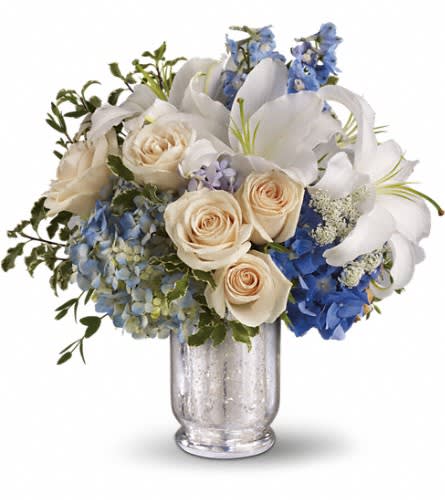 Teleflora's Seaside Centerpiece - Spend a peaceful afternoon at the seaside with this breathtaking centerpiece. Lush blue hydrangea and delphinium evoke sea and sky while creamy roses white lilies and delicate Queen Anne's lace whisper of sand and sea foam. Presented in an antiqued Mercury Glass Vase it's an elegant choice for weddings and showers. Two shades of blue hydrangea are mixed with crème roses light blue delphinium fragrant white oriental lilies delicate Queen Anne's lace parvifolia eucalyptus and green pitta negra in a beautiful Mercury Glass Antique Hurricane. Approximately 14 3/4" W x 14 3/4" H Orientation: All-Around As Shown : T184-1A