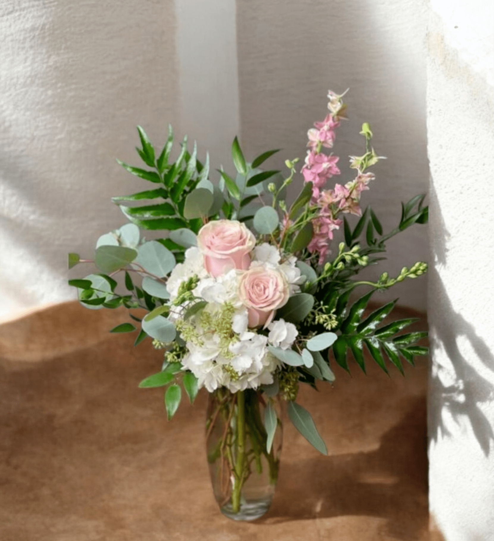 HOUSE BOUQUET HIDRANGEAS WITH ROSES - Soft pale pink roses nestled against clusters of white hydrangeas give this house bouquet a soft, romantic look perfect for an anniversary, birthday, or thoughtful thank-you gift. The arrangement reads like a fresh-cut garden with airy eucalyptus-like foliage and tall pink accent stems that lift the silhouette-she'll feel genuinely seen by the calm, celebratory mood it creates.  Delivered in a clear glass vase, the loose, asymmetrical styling lets stems and greenery extend outward for natural movement and texture. The palette of blush, white, lavender-tinged accents, and varied greens keeps the design light and elegant while remaining approachable for everyday gifting. Ideal for a loved one who prefers understated romance, the vase-ready arrangement arrives ready to display and enjoy.