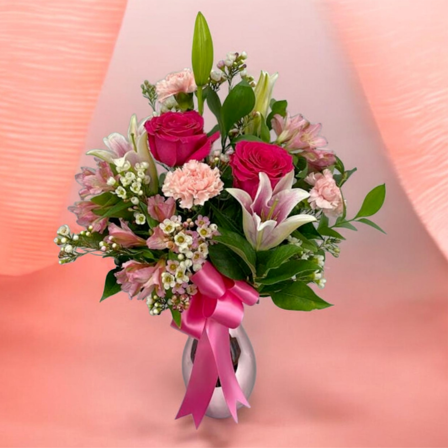 LILA MIX CARNATIONS - A lush mix of deep red roses and soft pink lilies and carnations comes together in LILA to create a romantic, polished bouquet for anniversaries, love and romance, or a thoughtful Mother's Day gift. The rich reds and blush pinks are balanced by crisp white blooms and fresh green foliage so the person you're thinking of will feel both celebrated and comforted - this arrangement also carries the formality suited to sympathy or a sincere "just because" moment.  Available in either a tall clear glass vase or a reflective silver vase, LILA pairs clustered red or vivid pink roses with airy white filler flowers, pale pink lilies, alstroemeria, carnations, and glossy greenery for a full, upright silhouette. The silver-vase option is finished with a bright pink satin ribbon for a gift-ready look; the clear-vase version showcases stems through the glass for a classic presentation. Perfect for birthdays, Valentine's Day, or as a funeral tribute, this arrangement arrives arranged and ready to display.